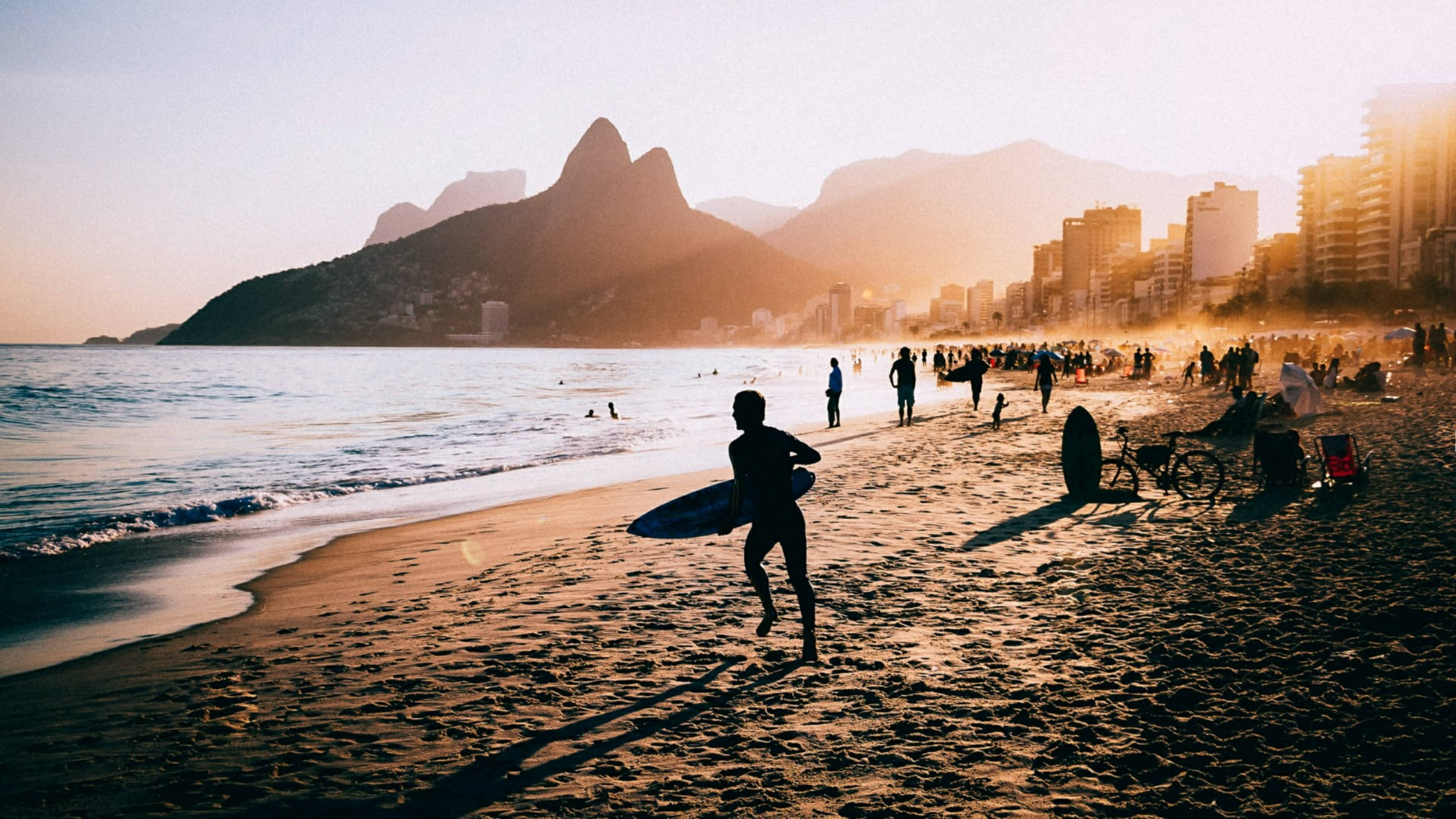 rio 桌布 壁紙 — 一名衝浪者在黃金時段沿著陽光明媚的海灘奔跑，背景是標誌性的山脈和城市天際線。