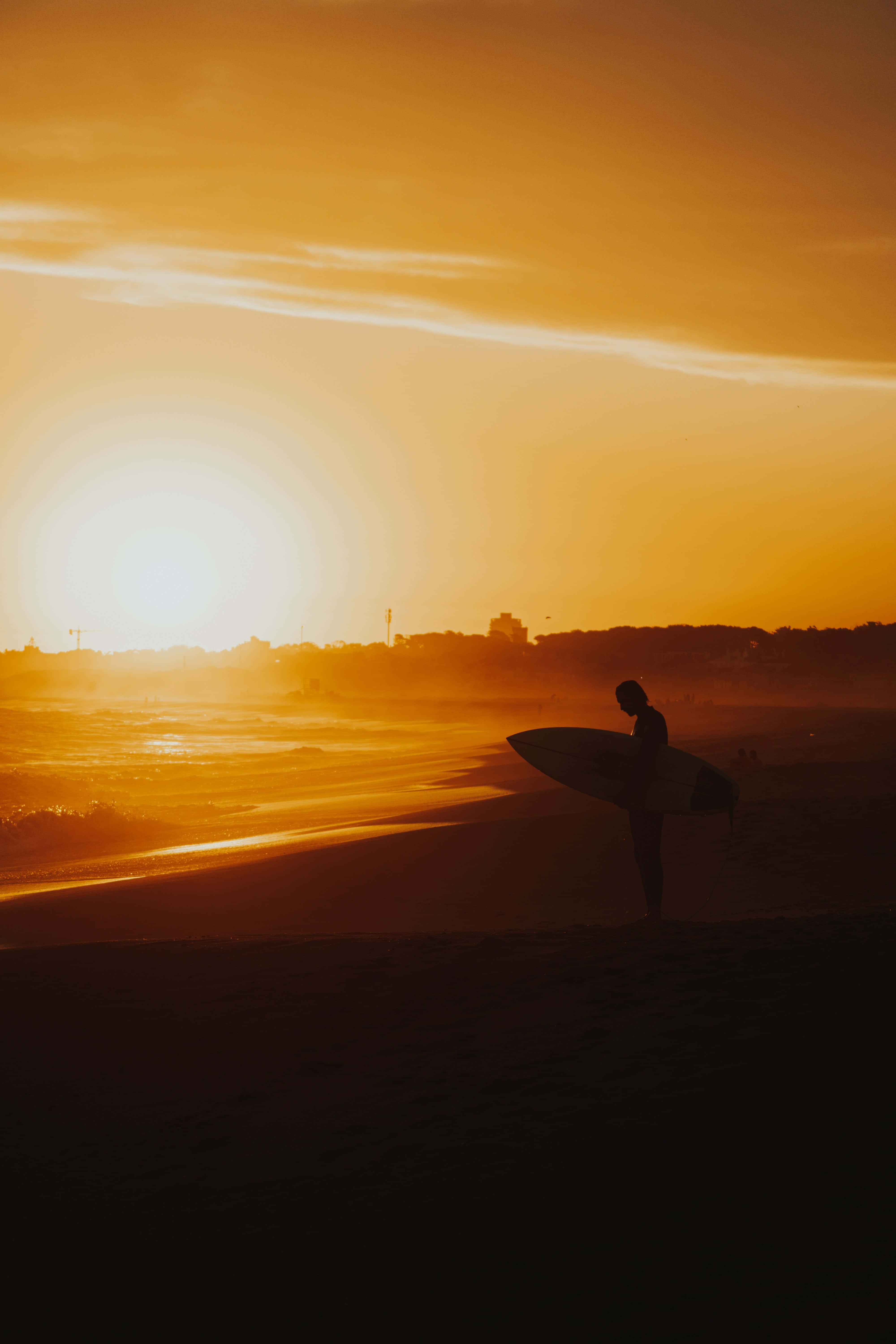 Aesthetic Beachy 桌布 壁紙 — 一名衝浪者的剪影站在日落時分的沙灘上，手持衝浪板，金色光芒灑滿地平線。