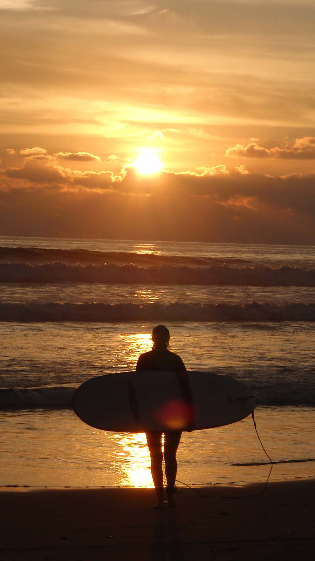 Surfing iPhone 桌布 壁紙 — 一名衝浪者站在沙灘上，手持衝浪板，在夕陽以火熱的橙色和金色色調沉入海洋時形成剪影。