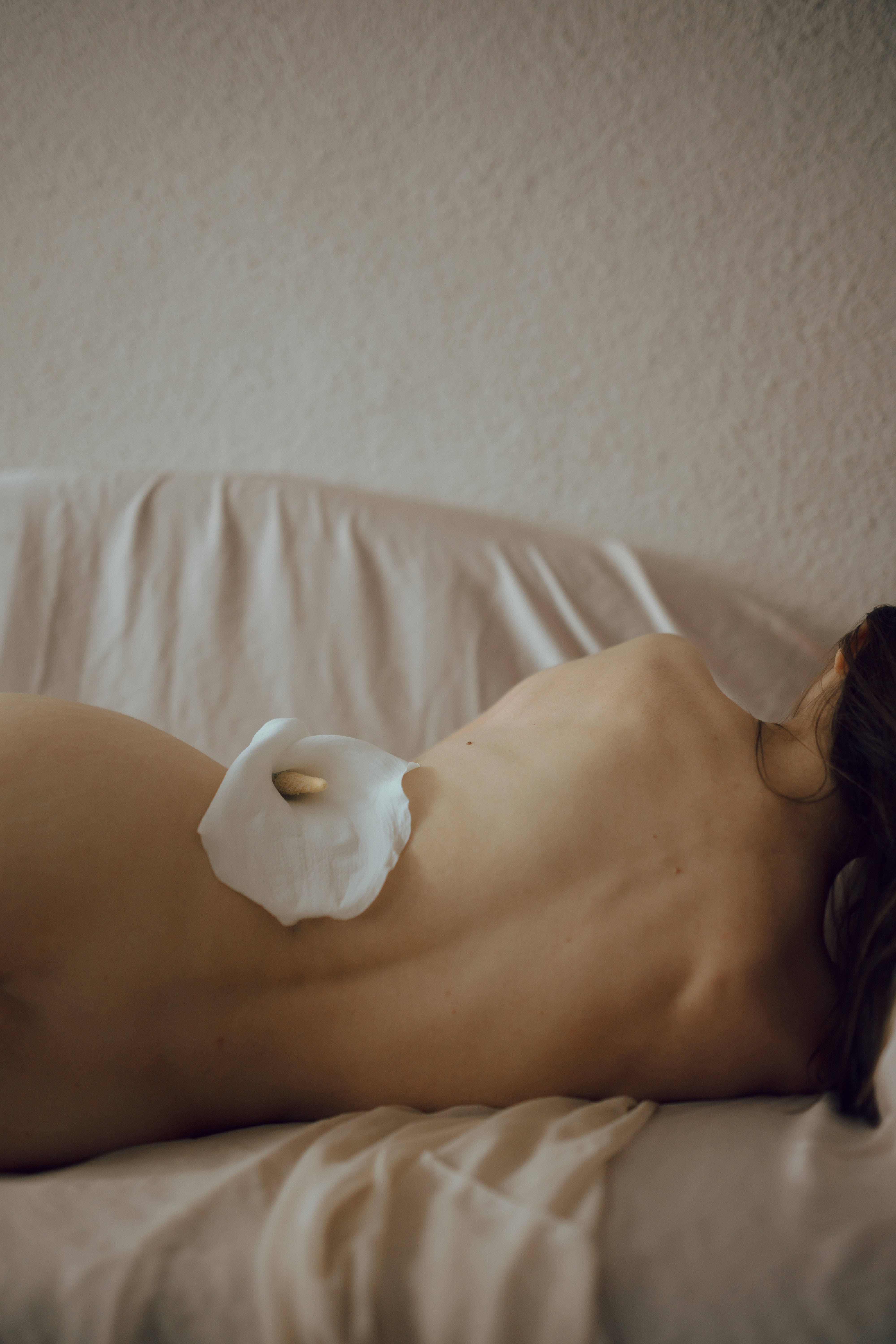 Sexy 桌布 壁紙 — 一名裸體女性側臥在床上，一朵白色馬蹄蓮放在她的背上，背景是帶有紋理的米色牆壁。