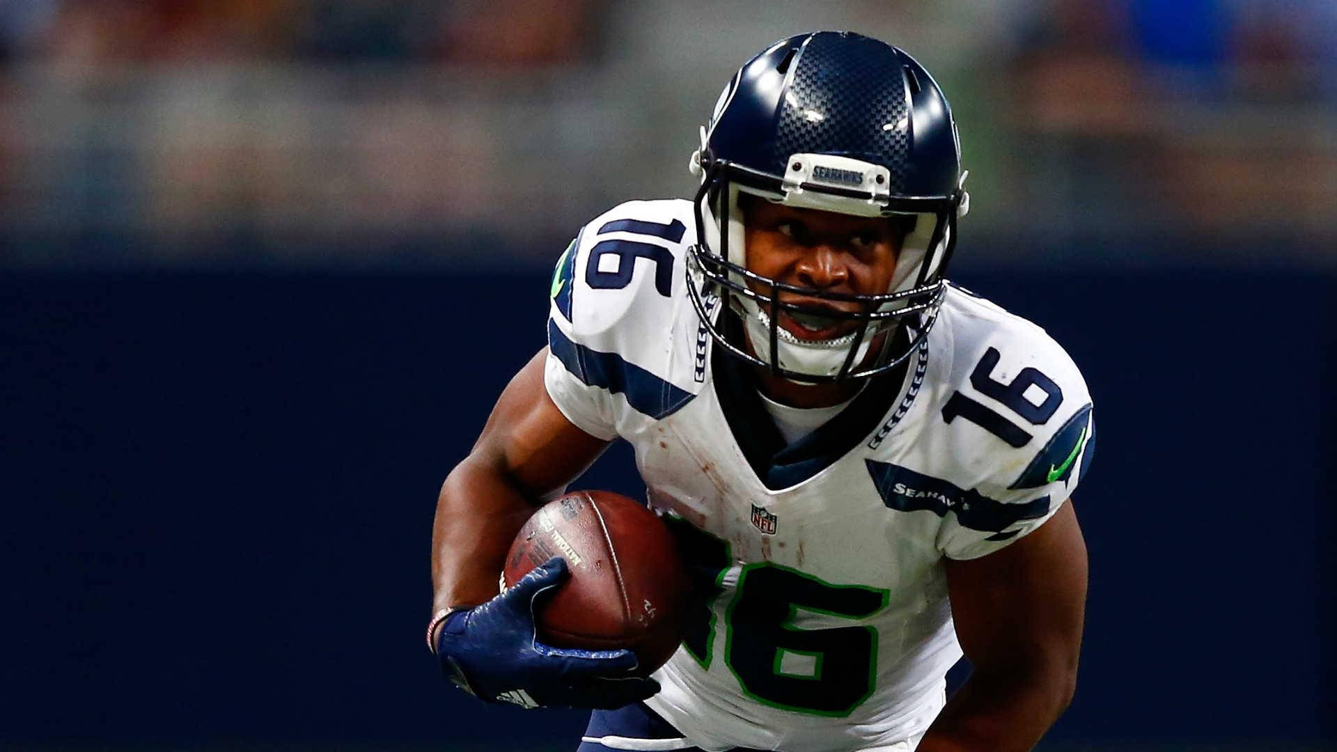 Seattle Seahawks 桌布 壁紙 — 一名西雅圖海鷹隊美式足球員身穿白色和藍色球衣，在球場燈光下比賽中帶球奔跑。