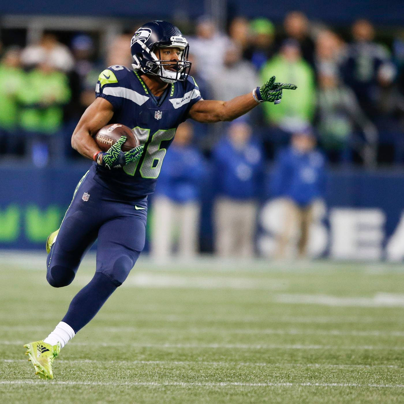Tyler Lockett 桌布 壁紙 — 一名西雅圖海鷹隊美式足球員身穿藍綠球衣，在夜間比賽的球場燈光下持球衝刺。