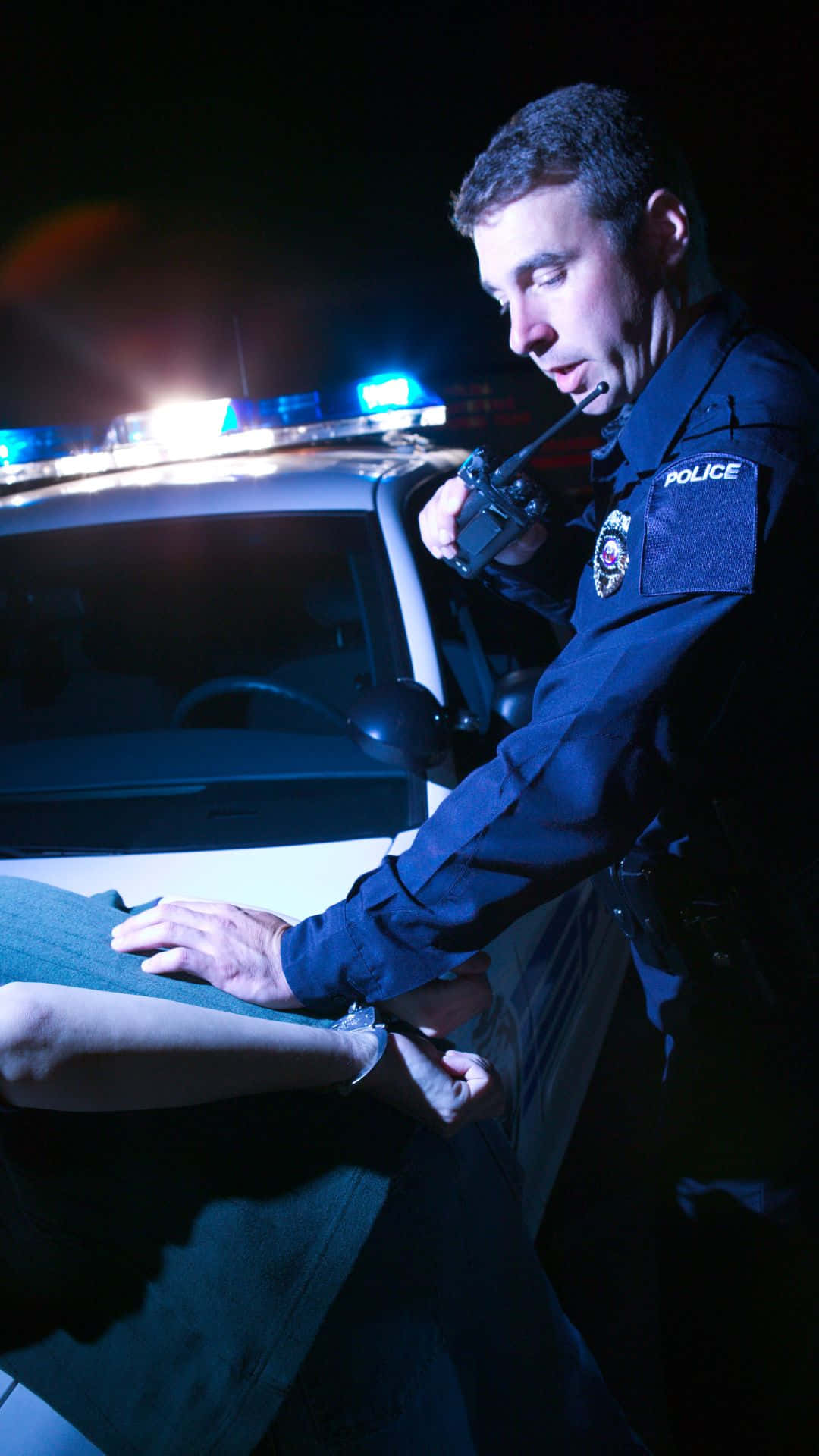 cop 桌布 壁紙 — 一名警察在夜間於一輛閃爍藍燈的巡邏車旁給一個人上手銬。