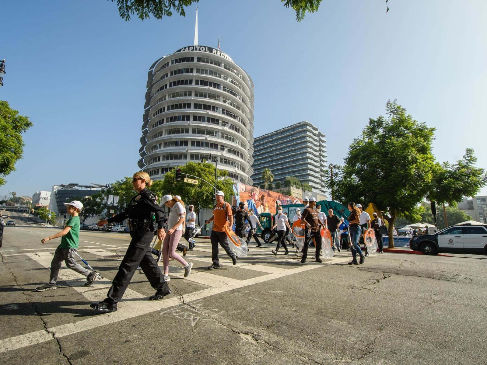 人物 桌布 壁紙 — 一名警察引導行人穿過陽光明媚的街道，前方是標誌性的 Capitol Records 大樓。