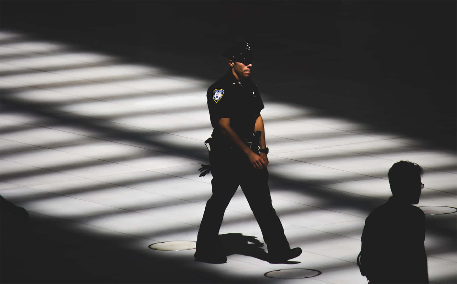 security 桌布 壁紙 — 一名警察走過瓷磚地板上強烈照明的條紋，而附近有一人站在陰影中。