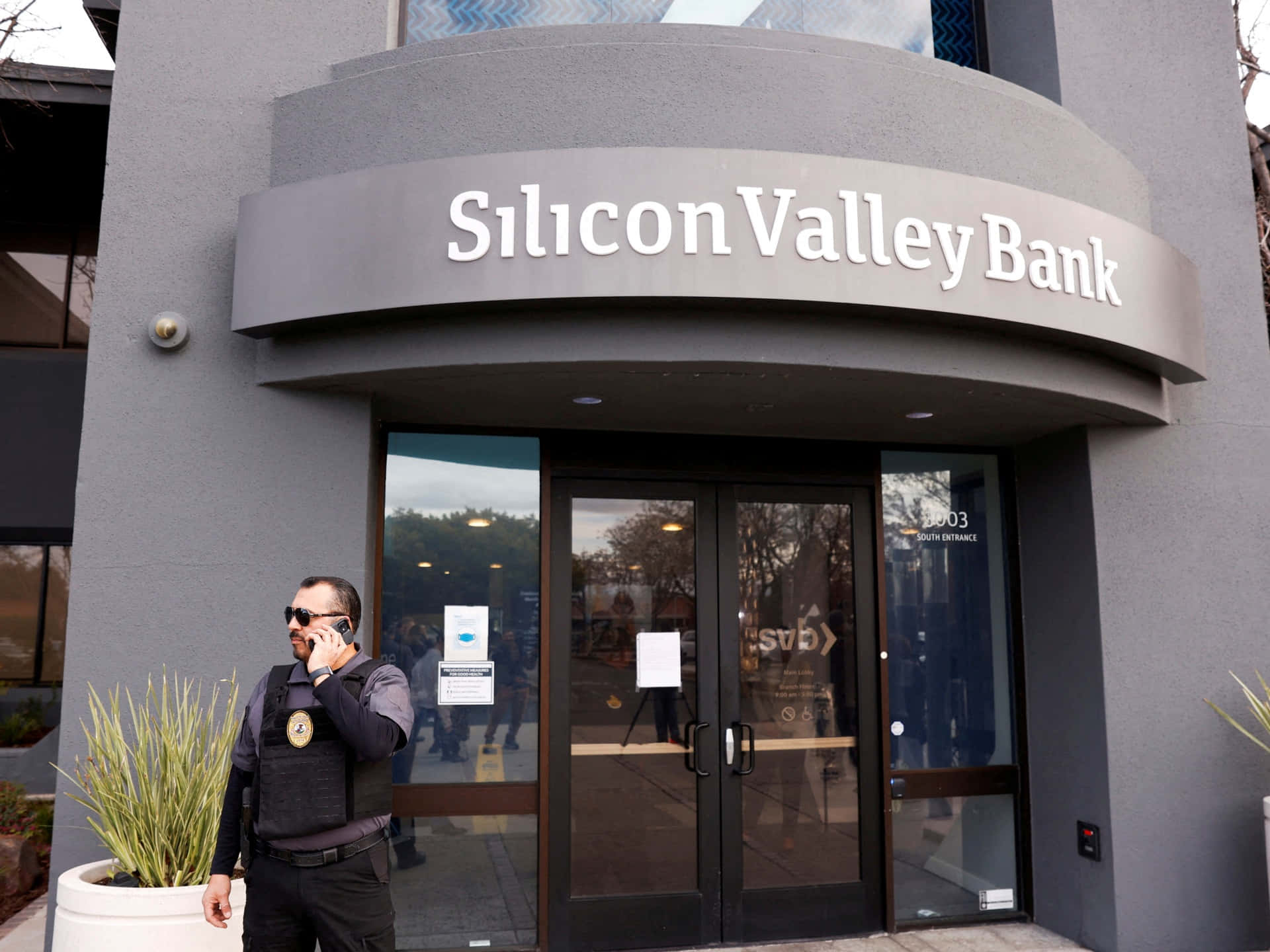 Bank 桌布 壁紙 — 一名警衛在矽谷銀行分行現代灰色外牆和玻璃門外講電話。