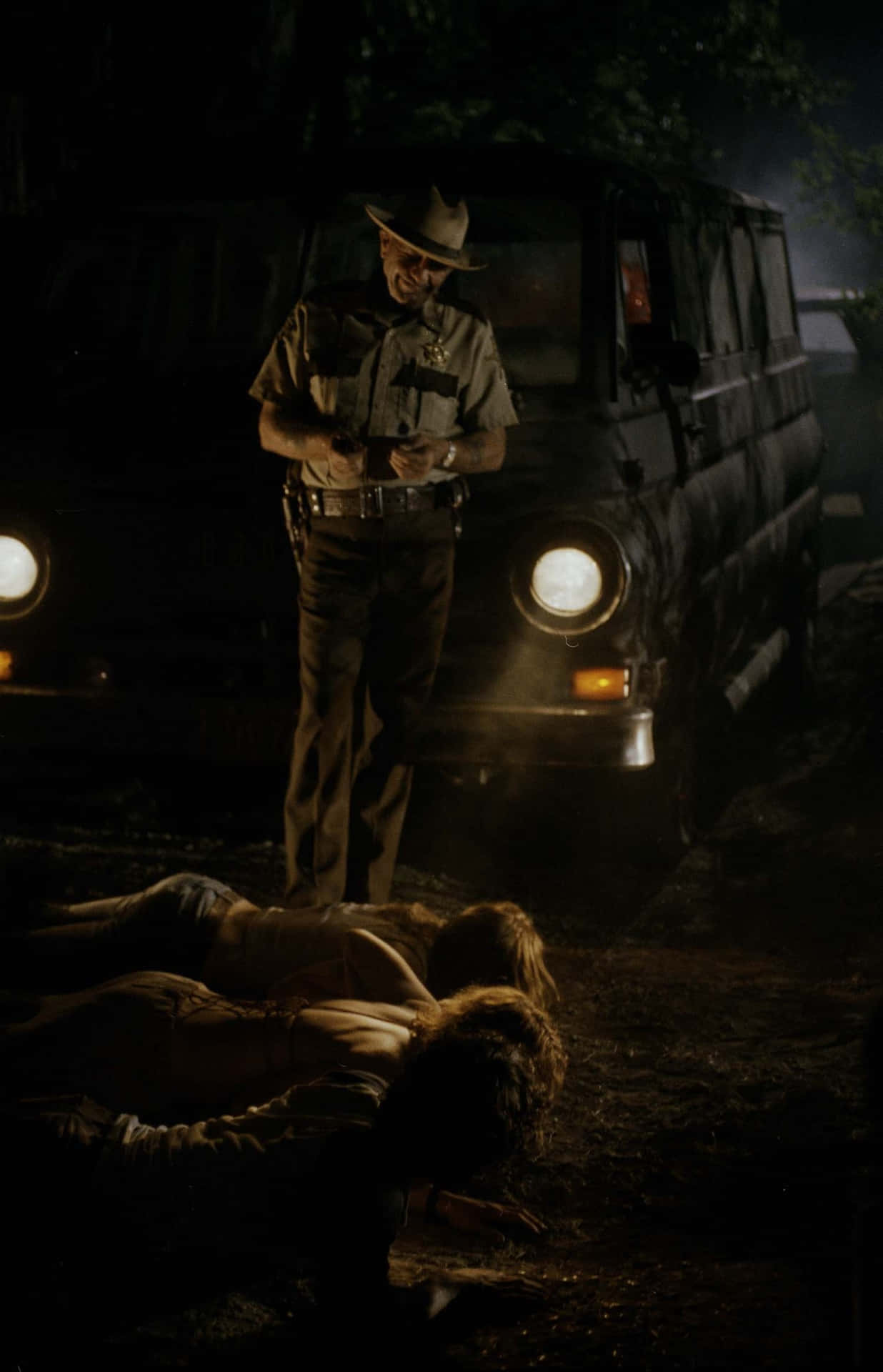 R. Lee Ermey 桌布 壁紙 — 一名警長在夜間站在兩個昏迷的人旁邊，旁邊有一輛開著車頭燈的貨車，位於黑暗的樹林地區。