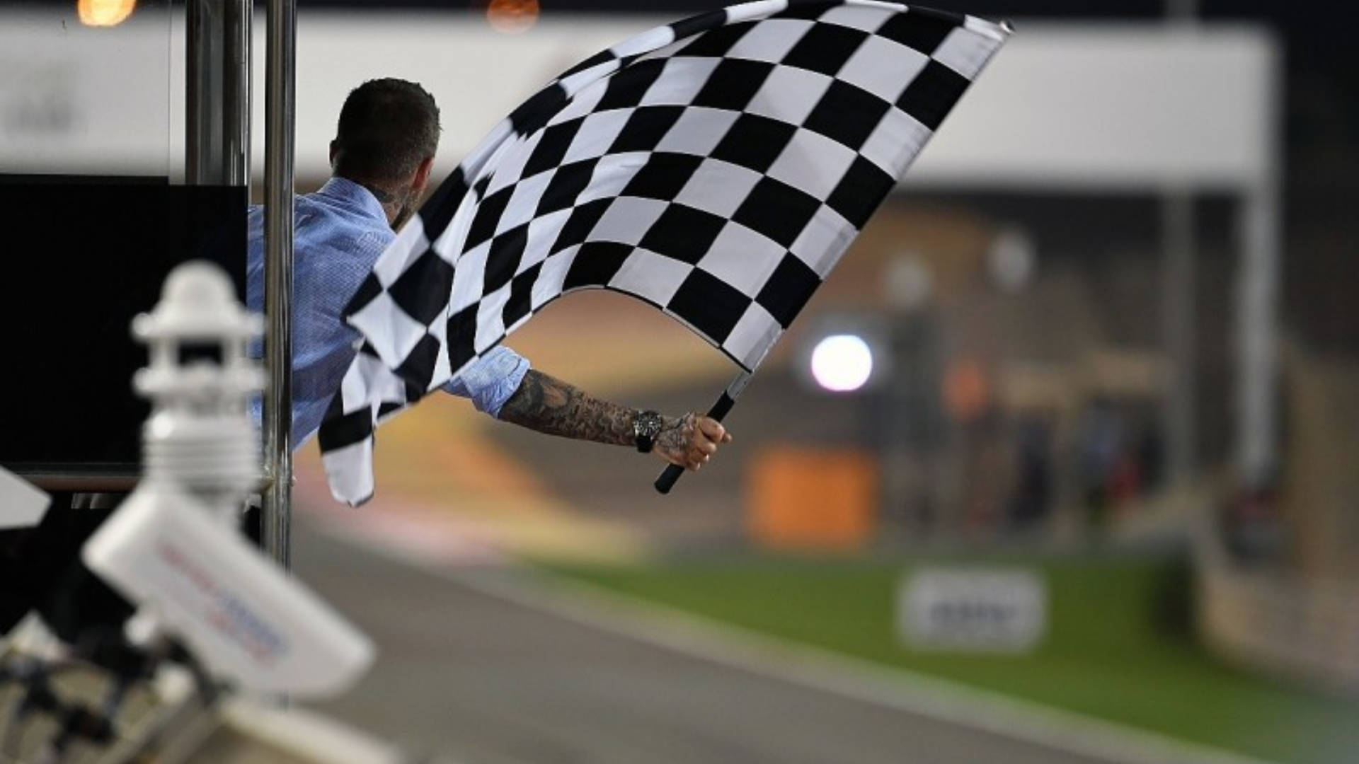 Checkered Flag 桌布 壁紙 — 一名賽事工作人員在夜間的賽車道上揮舞方格旗，背景有模糊的汽車和明亮的燈光。