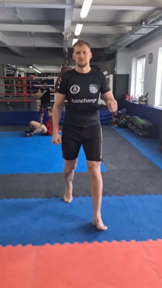 Athletic 桌布 壁紙 — 一名赤腳男子身穿黑色裝備，在拳擊館的藍色和紅色墊子上警覺地站立，背景中還有其他人在訓練。