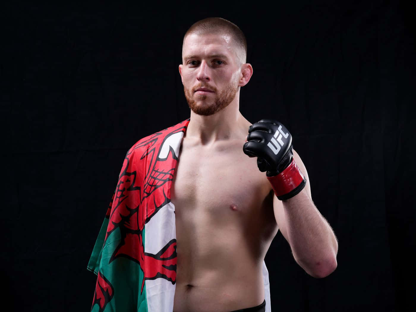 MMA 桌布 壁紙 — 一名赤膊、留鬍鬚的男性戰士，肩上披著威爾斯旗，在黑色背景前舉起UFC手套。