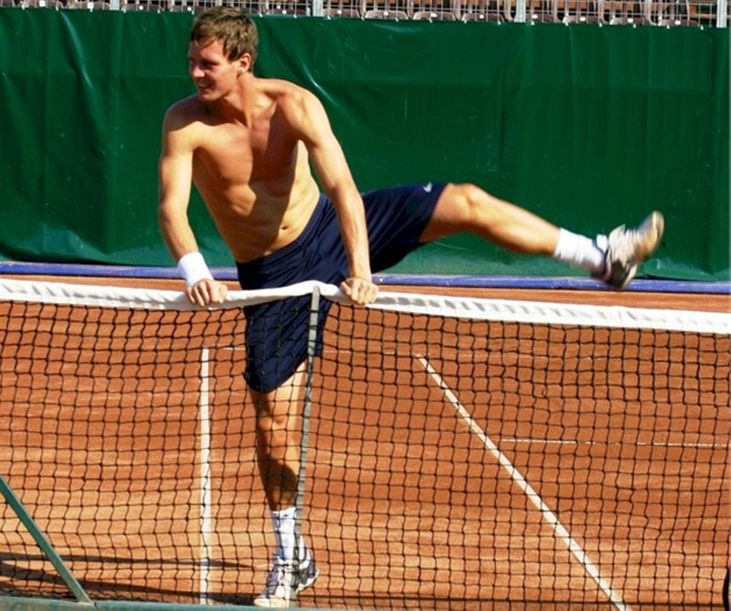 Tennis 桌布 壁紙 — 一名赤膊的男性網球選手在紅土球場上，一條腿伸展，臉上掛著笑容，身體傾斜在網上。