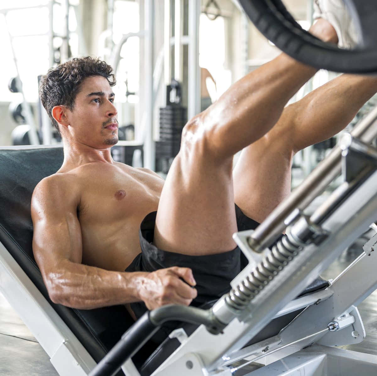 Exercise 桌布 壁紙 — 一名赤膊的肌肉男在明亮的健身房中，在腿部推舉機上進行劇烈運動。