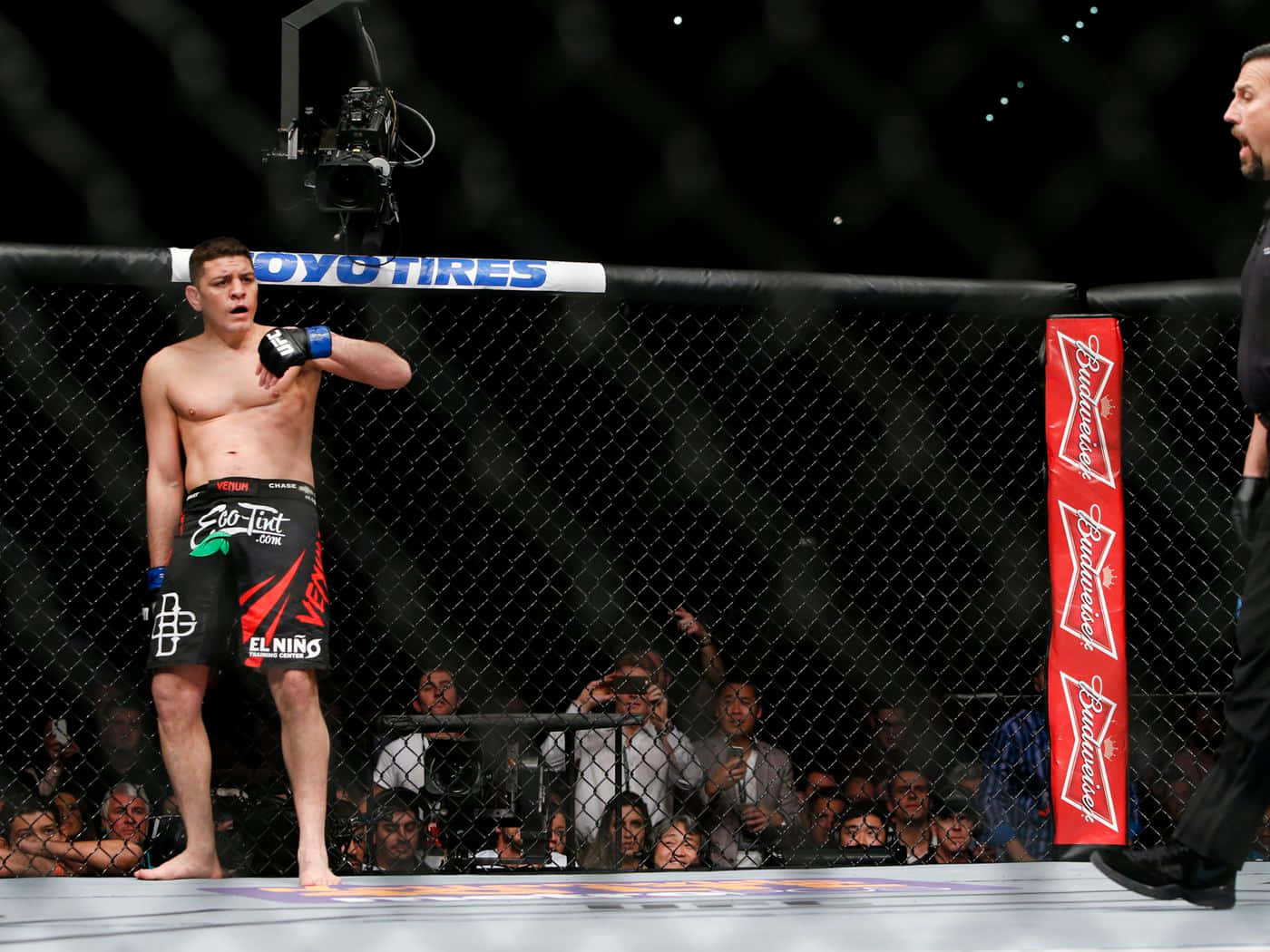 UFC 桌布 壁紙 — 一名赤裸上身的MMA拳手穿著黑色短褲，站在八角籠內，附近有裁判，觀眾在觀看。