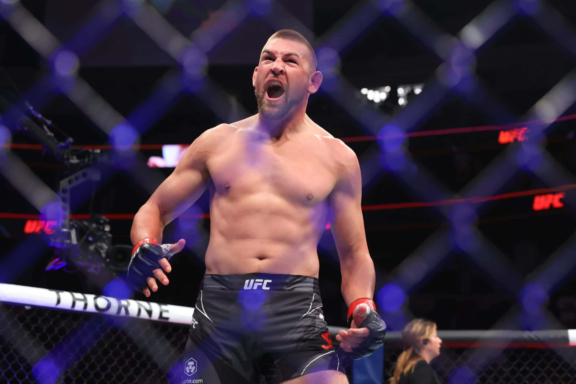 UFC 桌布 壁紙 — 一名赤裸上身的UFC拳手在八角籠內，在藍色燈光和模糊的籠子背景下，發出勝利的咆哮。