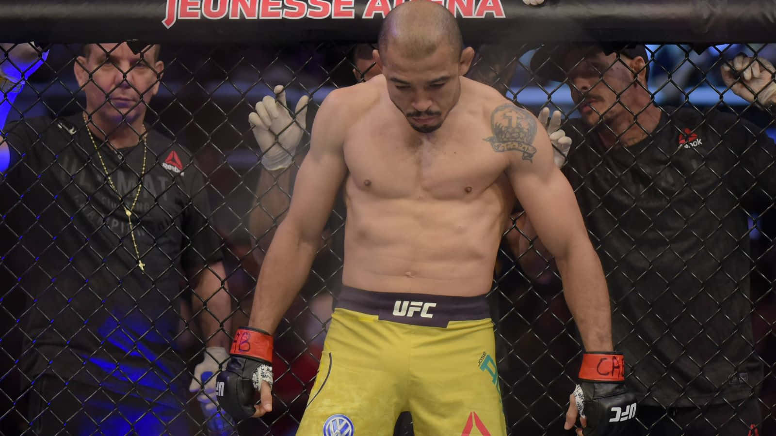 MMA 桌布 壁紙 — 一名赤裸上身的UFC拳手穿著黃色短褲，沮喪地站在八角籠內，附近有工作人員。