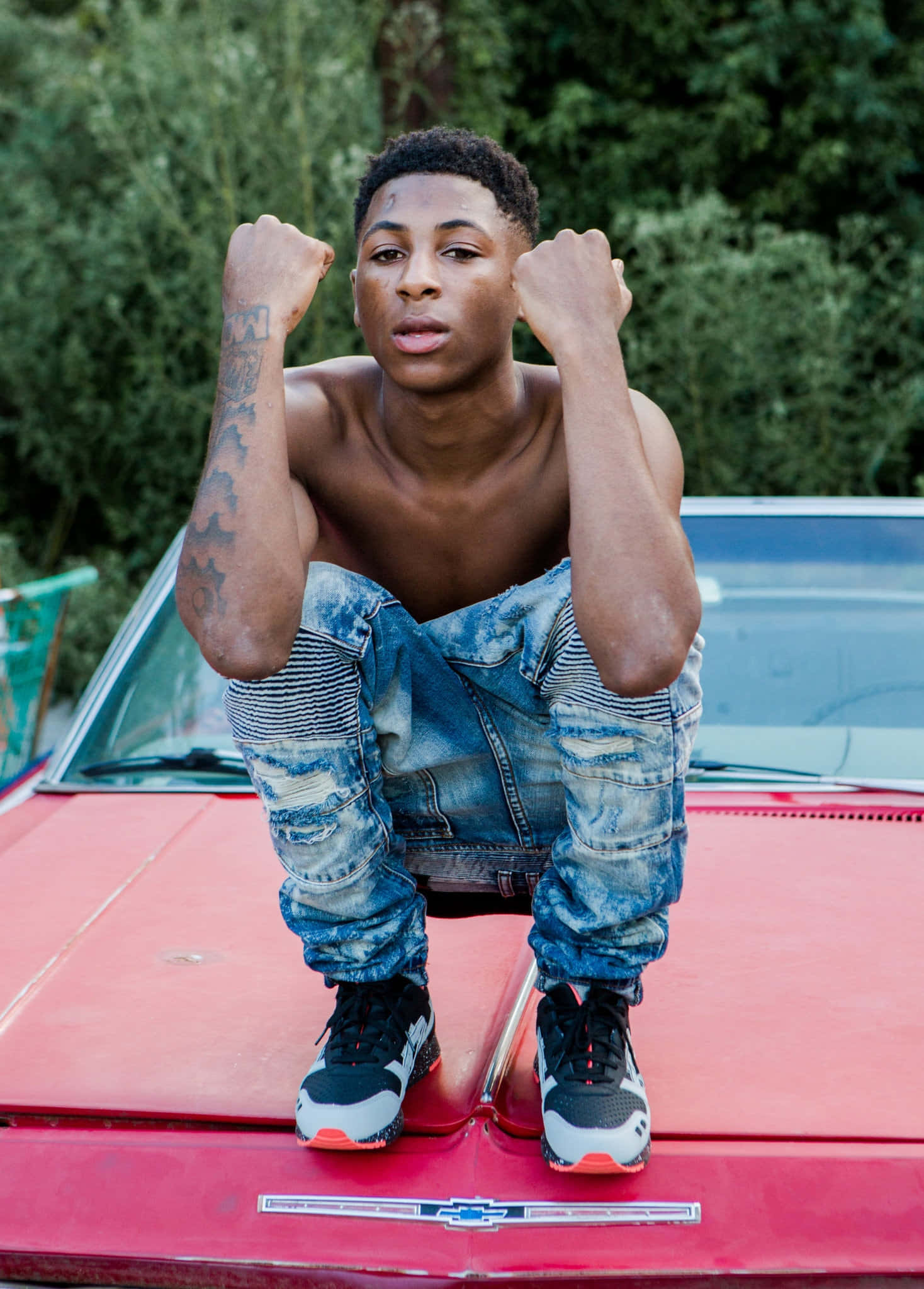 Youngboy 桌布 壁紙 — 一名赤裸上身的年輕刺青男子自信地蹲在紅色經典汽車的引擎蓋上，背景是茂密的綠色植物。