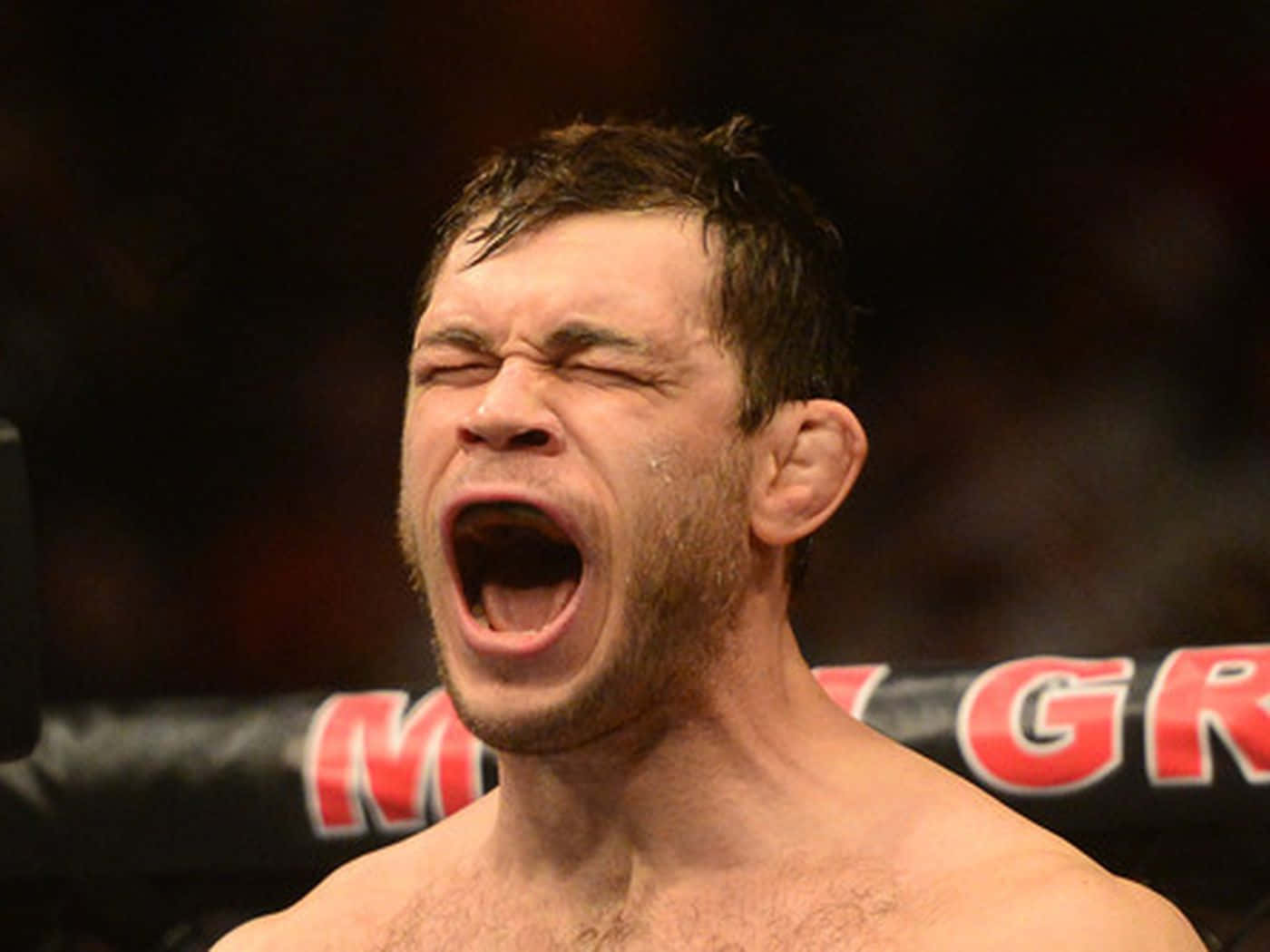 運動 桌布 壁紙 — 一名赤裸上身的戰士在MMA八角籠內勝利吶喊，背景有模糊的紅色品牌標誌。