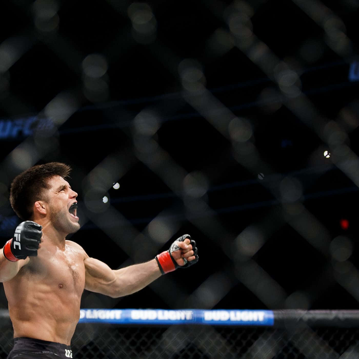 UFC 桌布 壁紙 — 一名赤裸上身的拳手在昏暗的UFC八角籠內高舉拳頭，發出勝利的咆哮。