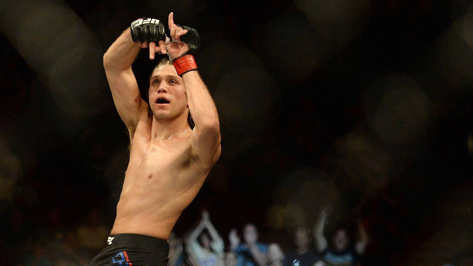 Brian Ortega 桌布 壁紙 — 一名赤裸上身的拳手在燈光昏暗的競技場內，模糊的觀眾背景下，勝利地舉起戴著手套的拳頭。