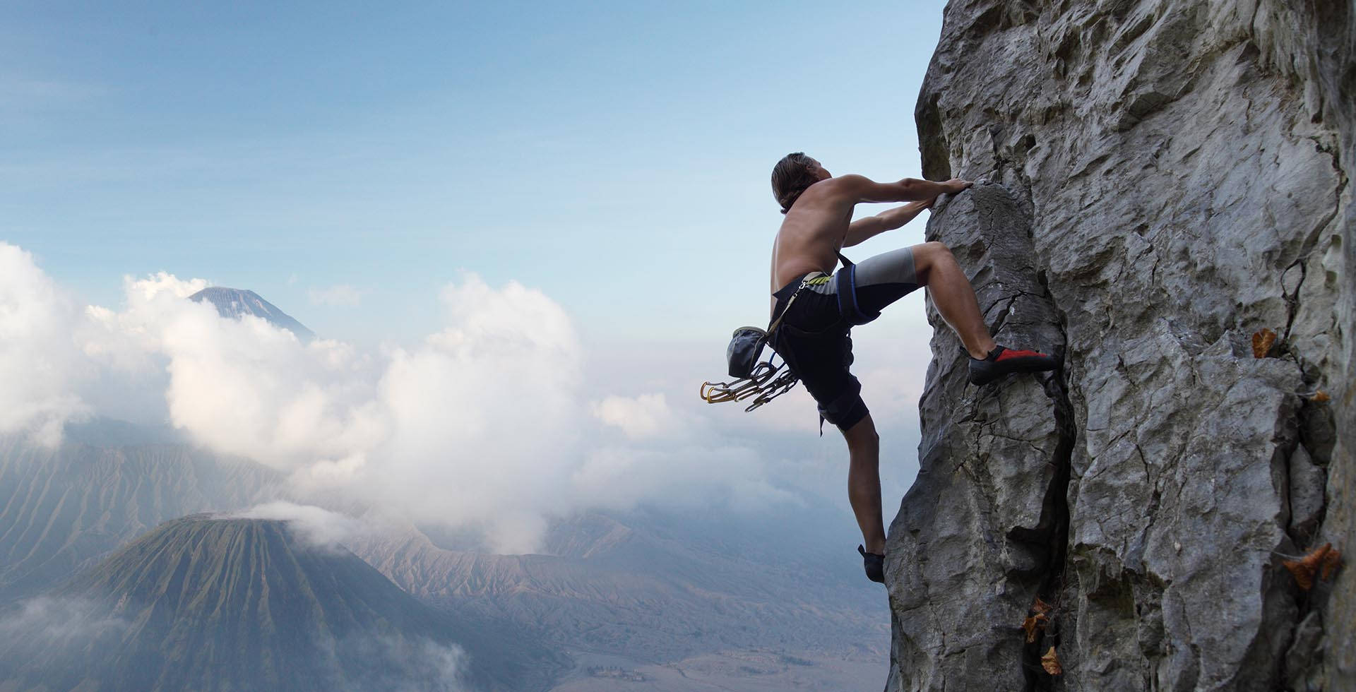 Climbing 桌布 壁紙 — 一名赤裸上身的攀岩者正在晴朗藍天下，攀登霧氣繚繞的火山峰頂之上的崎嶇懸崖面。