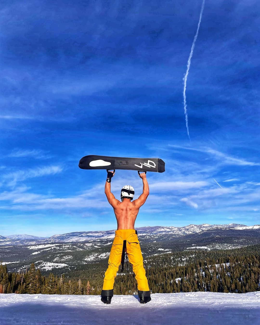 Ski Mountain 桌布 壁紙 — 一名赤裸上身的滑雪板運動員穿著鮮黃色褲子，在白雪覆蓋的山脊上高舉滑雪板，背景是帶有凝結尾跡的鮮豔藍天。