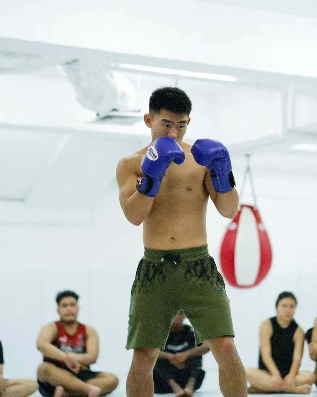 健身房 桌布 壁紙 — 一名赤裸上身的男子，戴著藍色拳擊手套和綠色短褲，在明亮的白色健身房內擺出戰鬥姿勢，背景有其他人坐著。