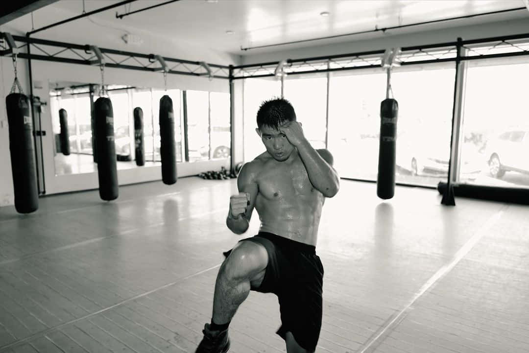 Athletic 桌布 壁紙 — 一名赤裸上身的男子在健身房內，於懸掛的重沙包和明亮的窗戶之間執行強力踢擊。