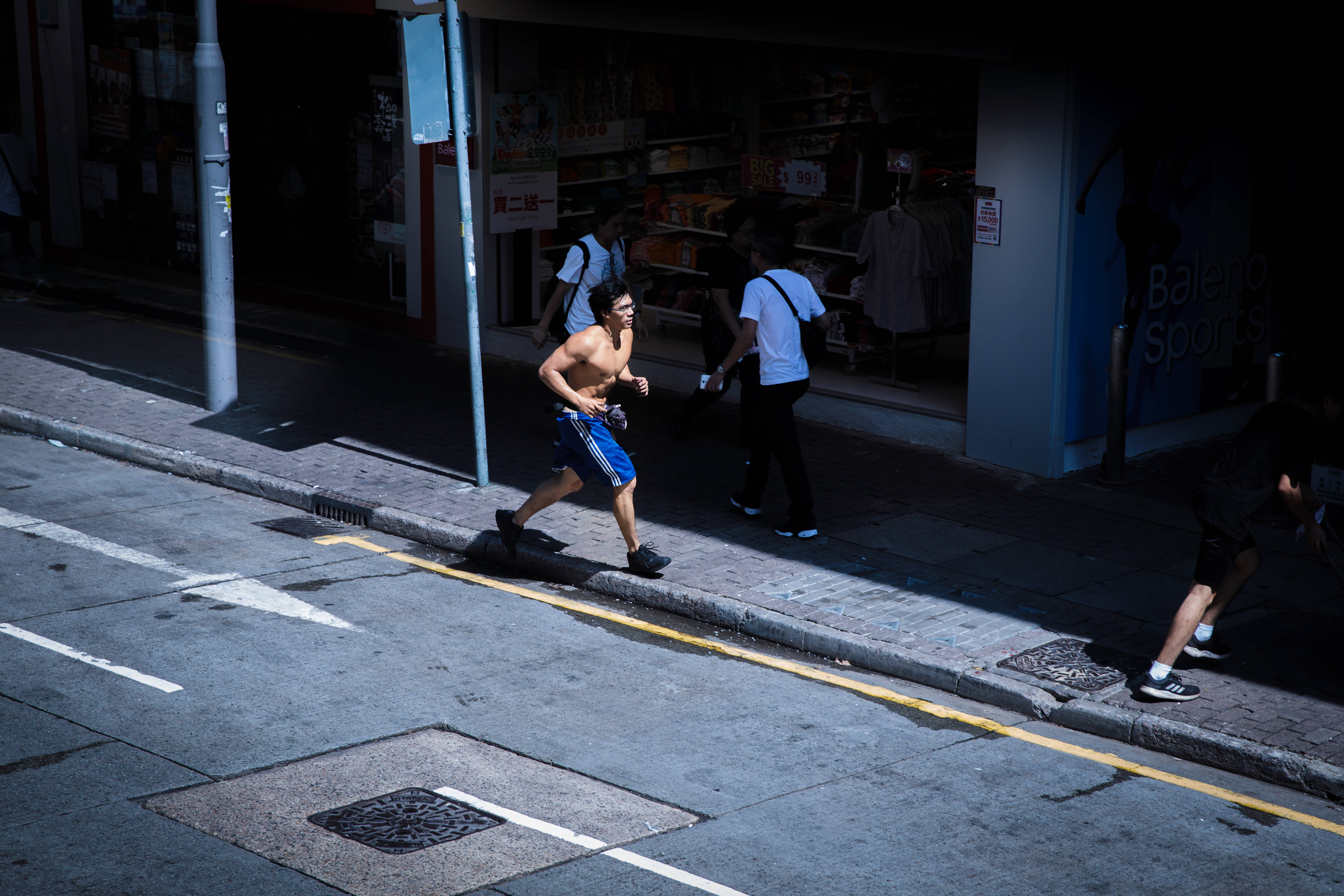 跑步者 桌布 壁紙 — 一名赤裸上身的男子在陽光明媚的城市人行道上奔跑，行人走過繁忙街道上的商店櫥窗。