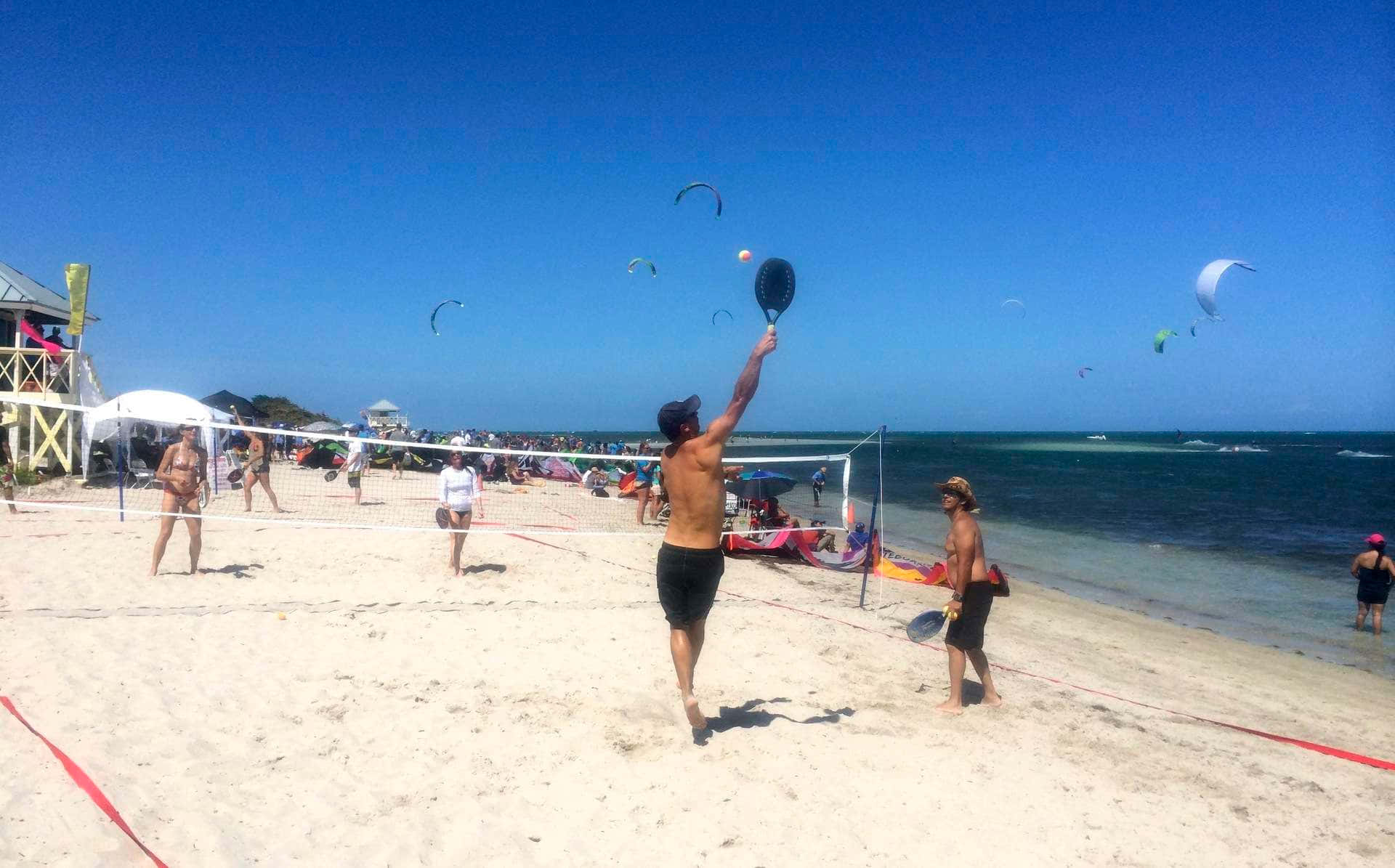 Beach Tennis 桌布 壁紙 — 一名赤裸上身的男子在陽光明媚的海灘上跳起擊打排球，而風箏衝浪者在清澈藍天下滑過碧綠的海洋。