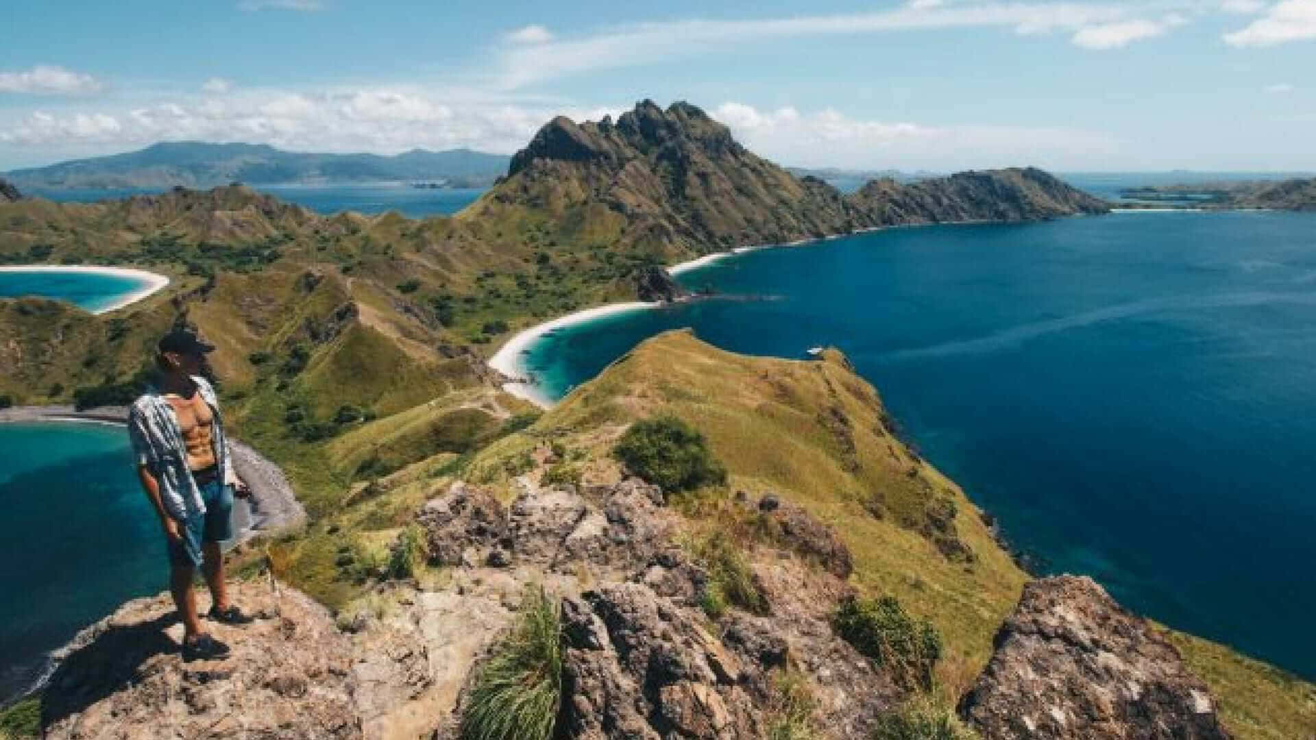 島嶼 桌布 壁紙 — 一名赤裸上身的男子站在岩石峭壁頂端，俯瞰著碧藍的潟湖和翠綠的山丘，背景是明亮的藍天。