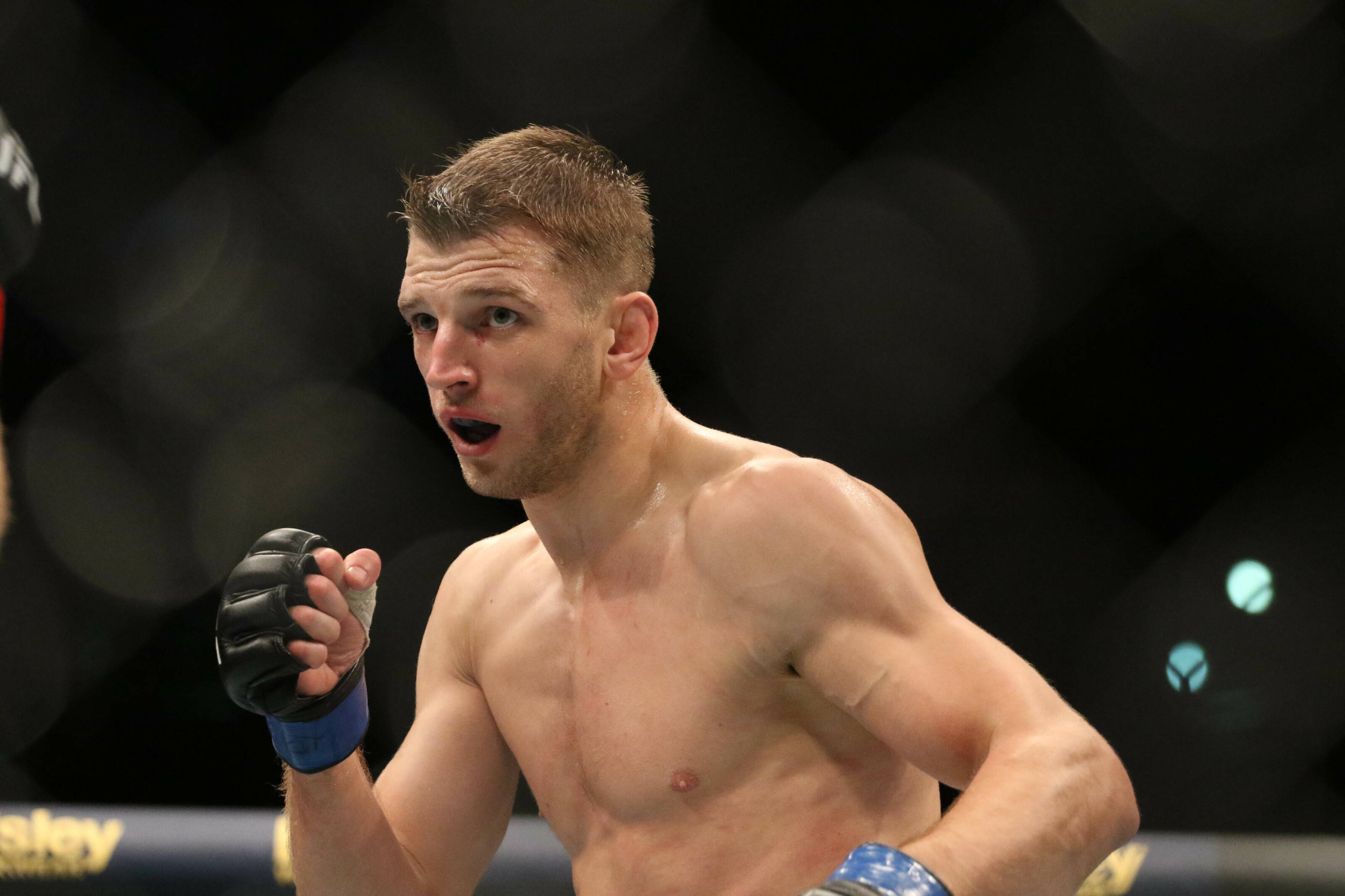 UFC 桌布 壁紙 — 一名赤裸上身的男性拳手，流著鼻血，表情激烈，在昏暗的八角籠中進行綜合格鬥比賽時大聲呼喊。