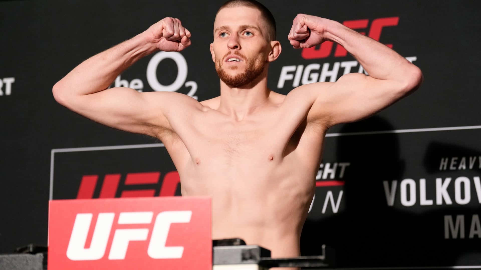 MMA 桌布 壁紙 — 一名赤裸上身的男性拳手在UFC稱重活動中展示肌肉，背景和體重計帶有品牌標誌。