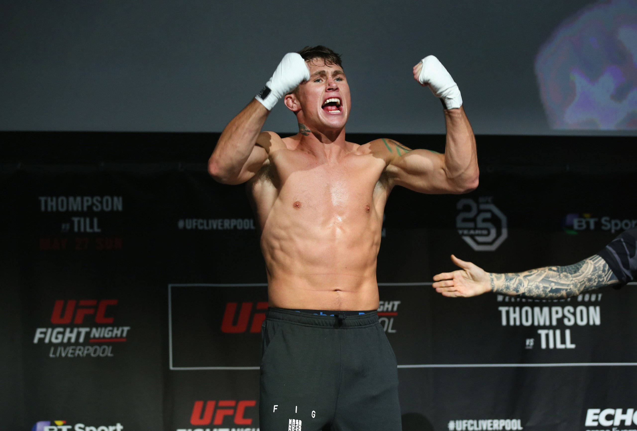 MMA 桌布 壁紙 — 一名赤裸上身的男性拳手在明亮的場館燈光下，於UFC稱重活動中舉起拳頭，發出勝利的怒吼。