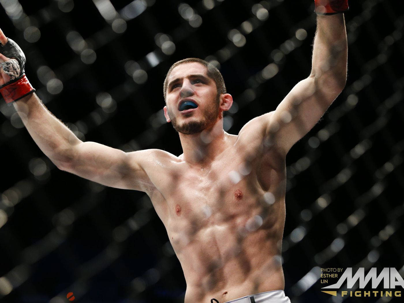 Islam Makhachev 桌布 壁紙 — 一名赤裸上身的男性拳手在昏暗的MMA八角籠內，背景是模糊的鐵絲網，他勝利地高舉雙臂。