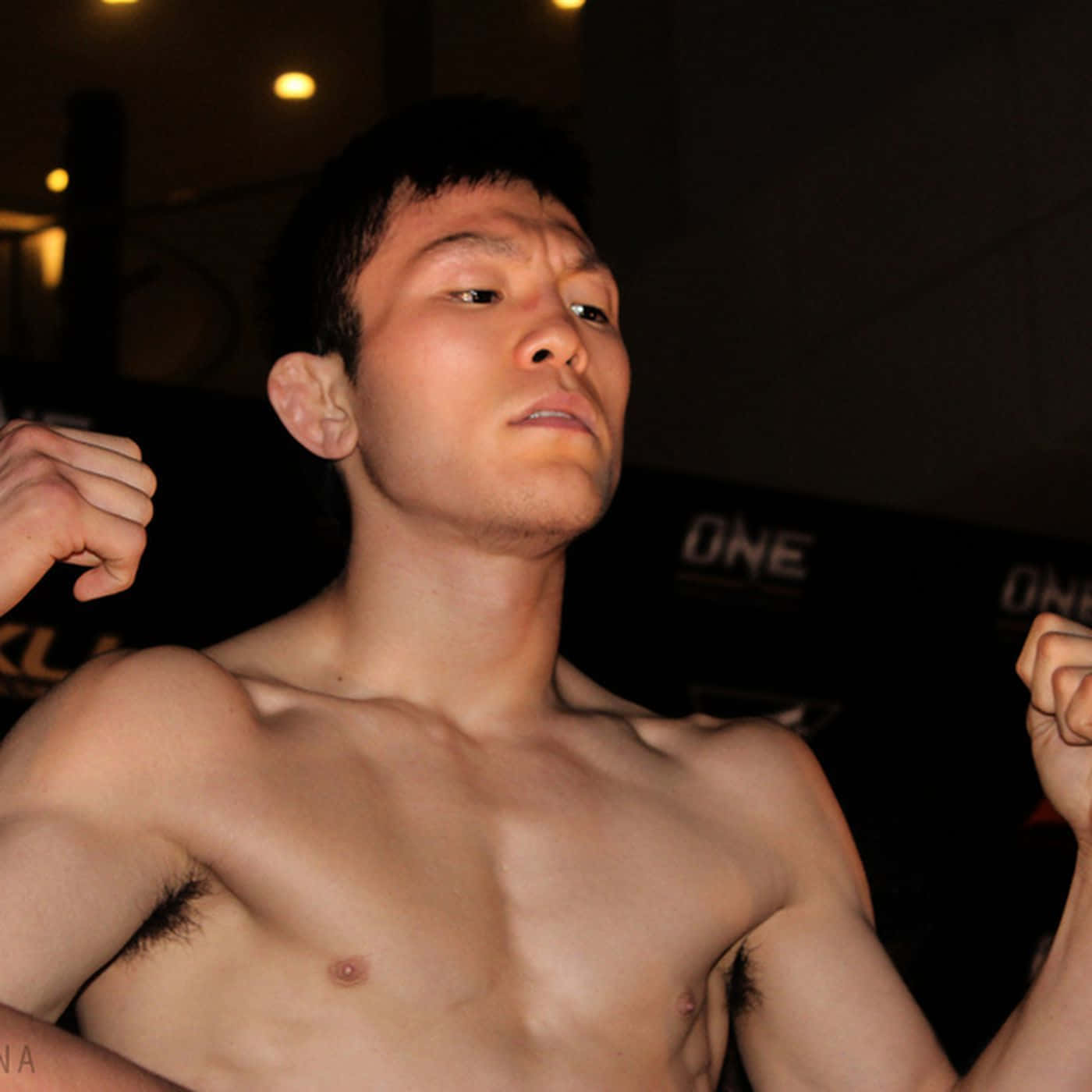 martial arts 桌布 壁紙 — 一名赤裸上身的男性拳手握緊拳頭，在昏暗的競技場中，背後有「ONE」品牌標誌，眼神專注。