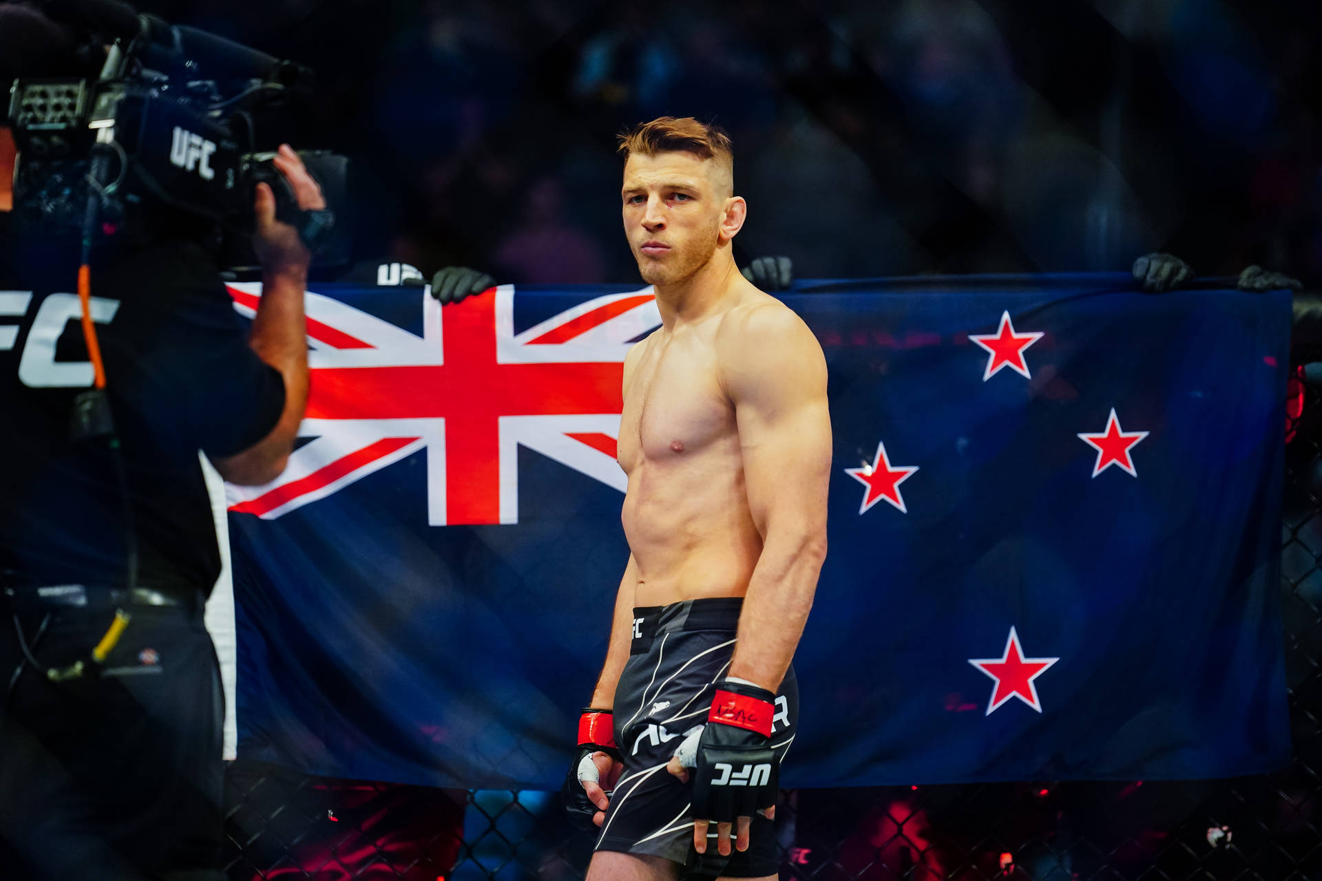 UFC 桌布 壁紙 — 一名赤裸上身的男性拳手站在籠中，手持紐西蘭國旗，攝製組正在拍攝他。