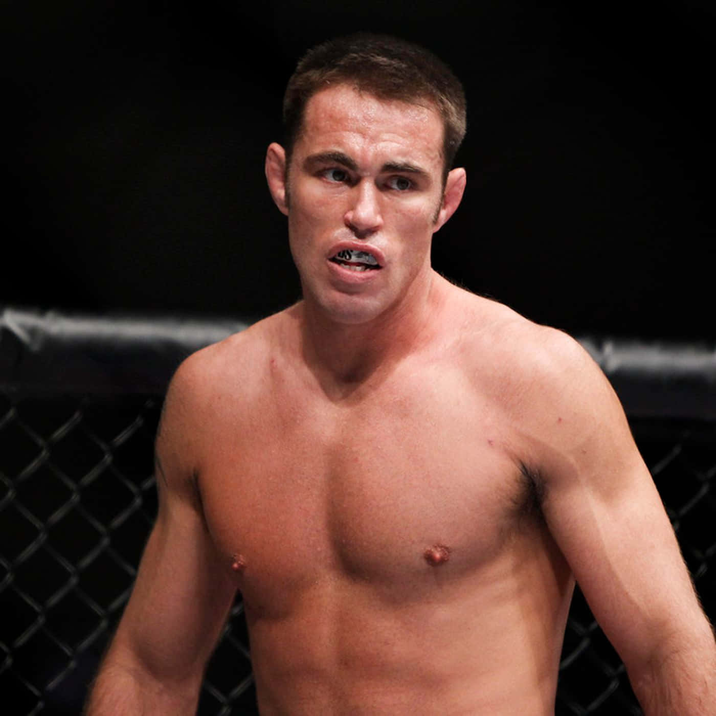 Athletic 桌布 壁紙 — 一名赤裸上身的男拳手，帶著強烈的專注力，站在燈光昏暗的MMA八角籠內。