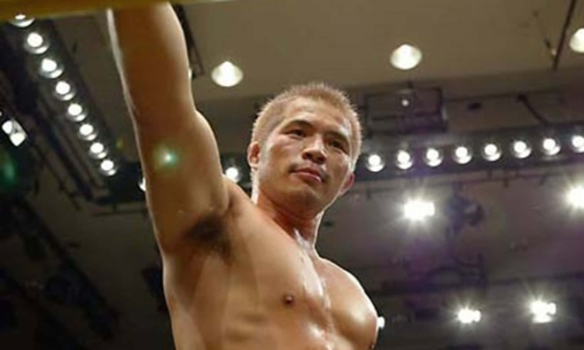 MMA 桌布 壁紙 — 一名赤裸上身的肌肉戰士在明亮的競技場燈光下勝利地舉起手臂。