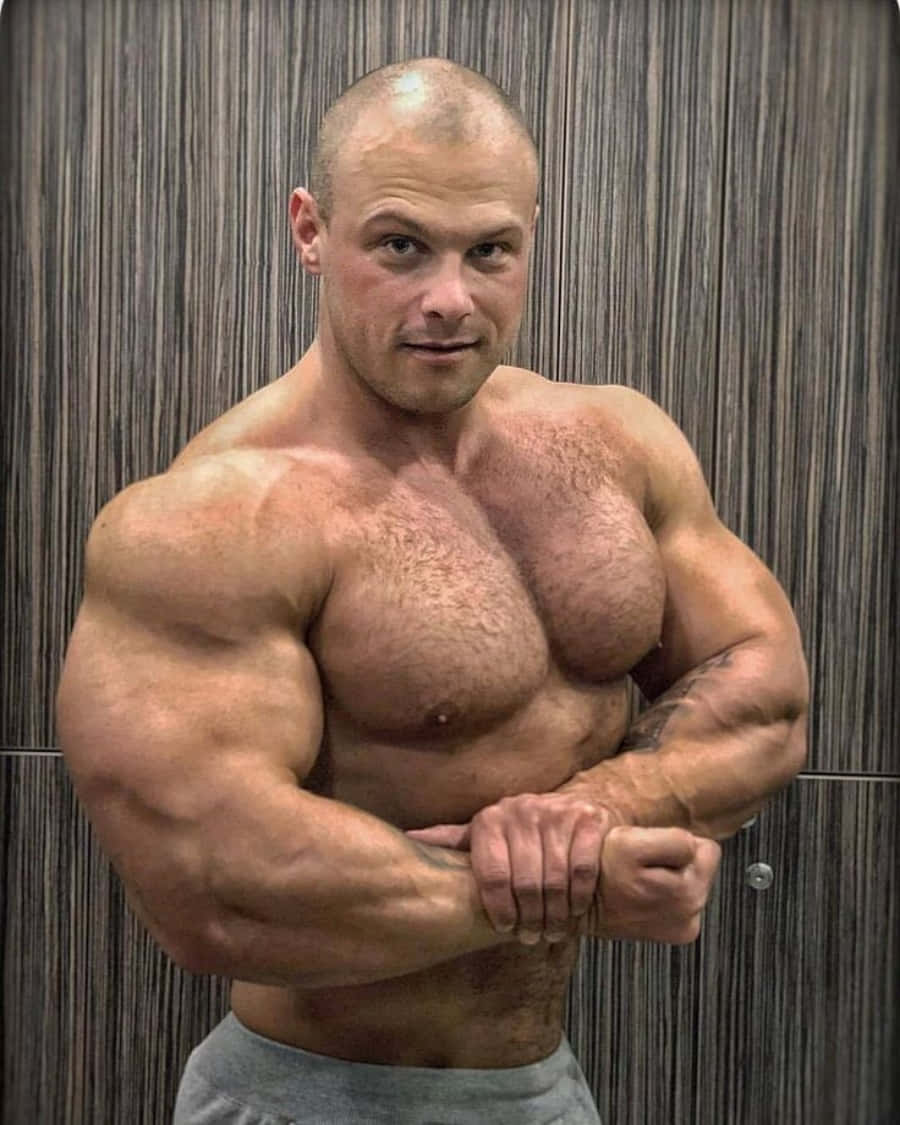 Muscle 桌布 壁紙 — 一名赤裸上身的肌肉男，在垂直的木鑲板牆前用力彎曲手臂，神情專注。