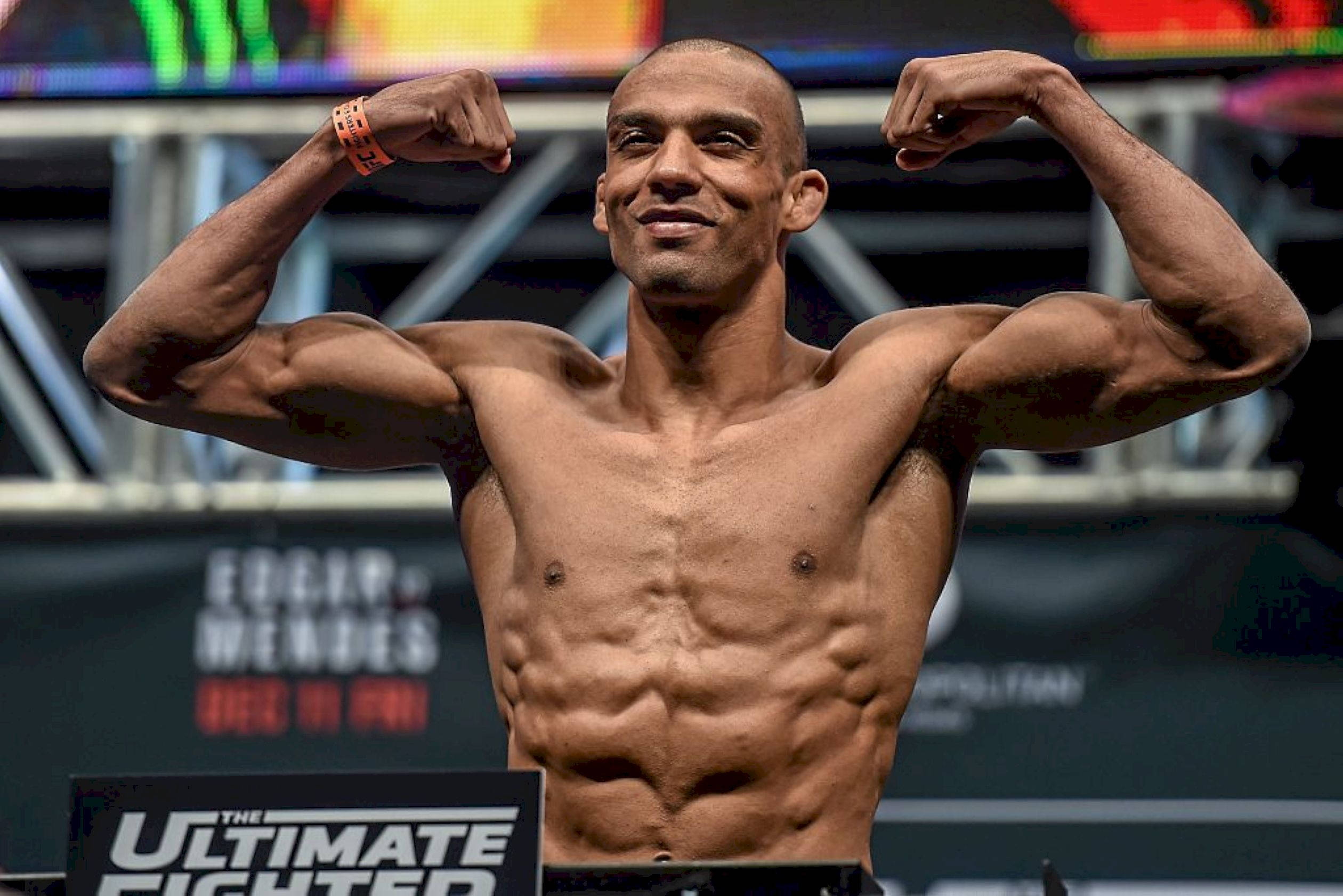 MMA 桌布 壁紙 — 一名赤裸上身的肌肉發達戰士在帶有「The Ultimate Fighter」品牌標誌的舞台上自信微笑，彎曲雙臂。