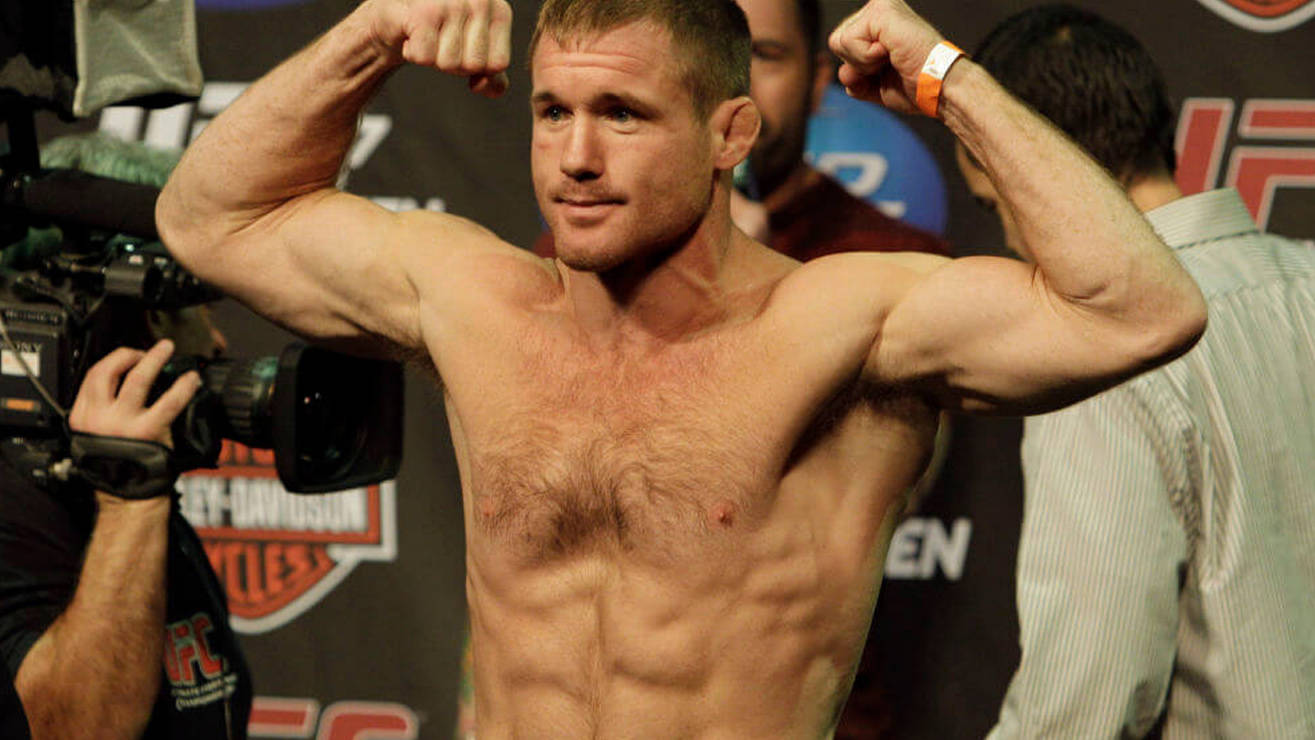 UFC 桌布 壁紙 — 一名赤裸上身的肌肉發達拳手在稱重活動中彎曲雙臂，背景有相機和UFC品牌標誌。