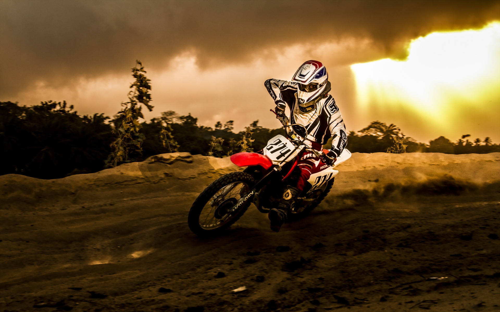 Motocross 桌布 壁紙 — 一名越野摩托車手在泥地賽道上傾斜入彎，金色陽光從背後的暴風雲中穿透而出。