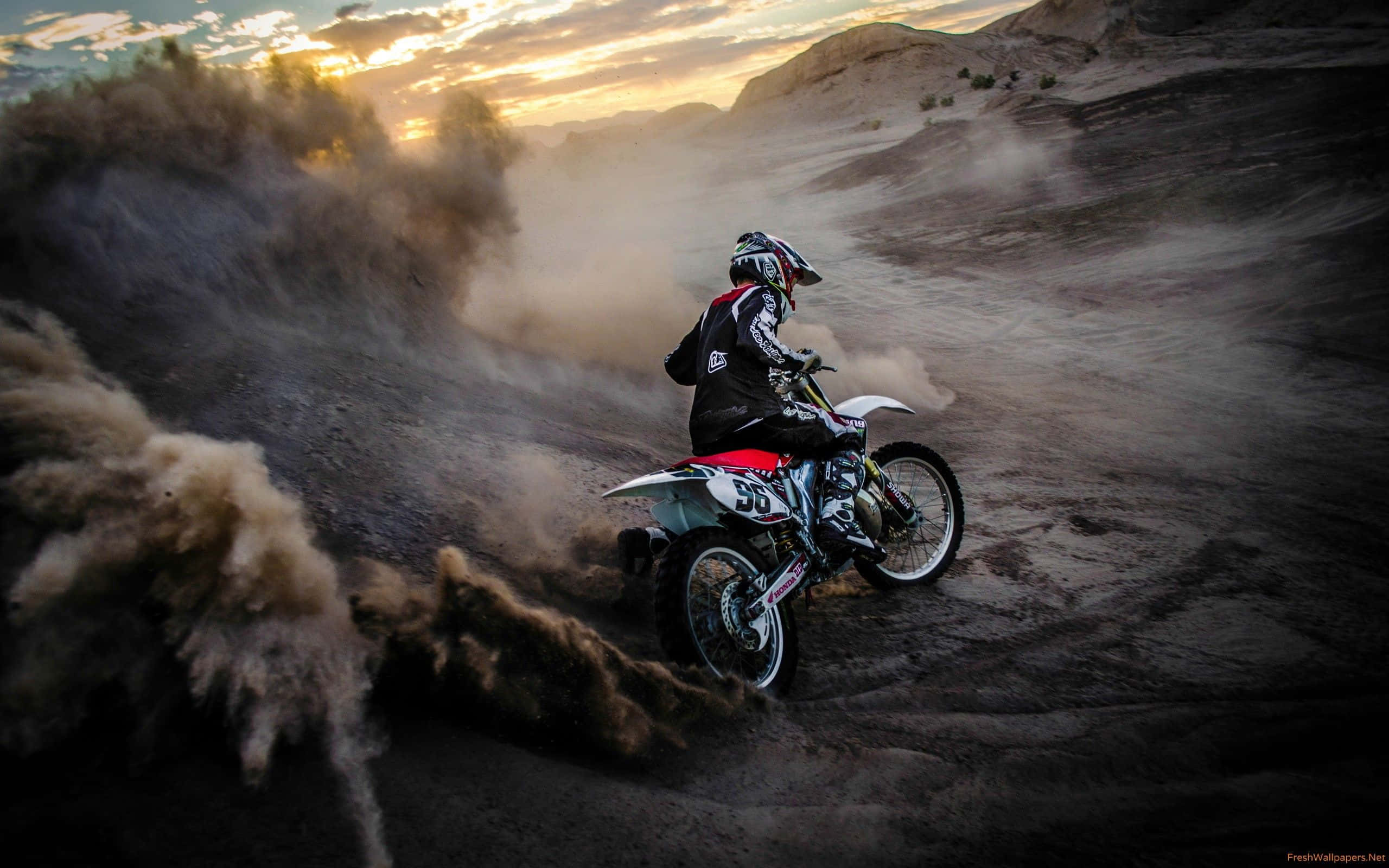 Honda Dirt Bike 桌布 壁紙 — 一名越野摩托車手在金色日落時分，在崎嶇的泥地上揚起塵土。