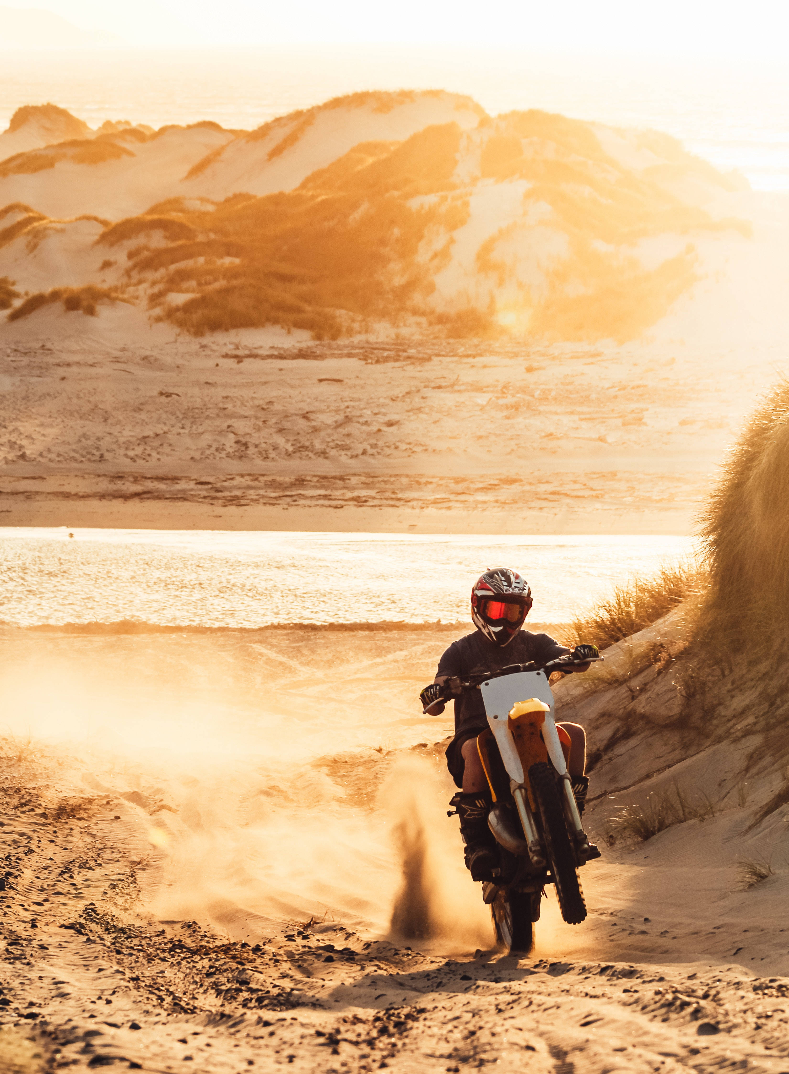 Dirtbike 桌布 壁紙 — 一名越野摩托車手在金色日落時分的沙丘上揚起沙塵，下方海浪拍打。