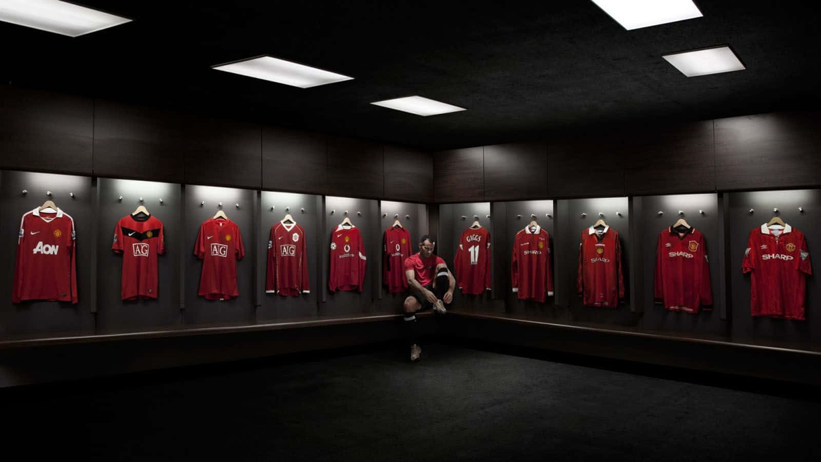 Manchester United Team 桌布 壁紙 — 一名足球員在昏暗的更衣室中跪下，周圍是掛在深色木牆上的一排排紅色曼聯球衣。