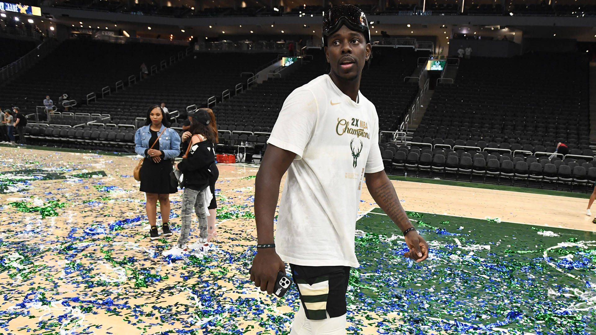 Jrue Holiday 桌布 壁紙 — 一名身穿 '2x NBA Champions' 球衣的籃球運動員在空無一人的看台後方，走過灑滿彩紙的球場。
