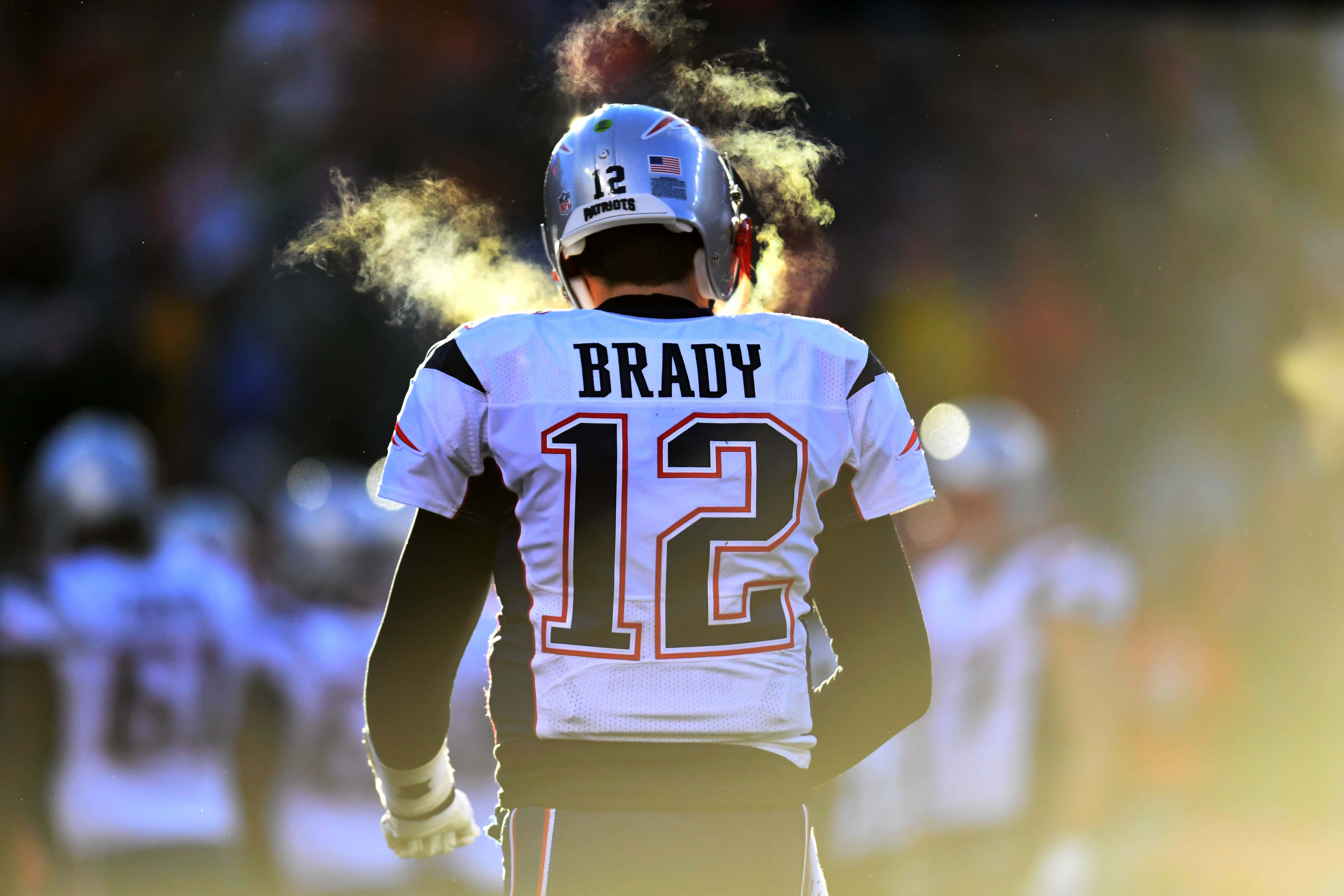 Tom Brady Goat 桌布 壁紙 — 一名身穿12號球衣和頭盔的美式足球員站在明亮的球場上，煙霧在他頭頂盤旋。
