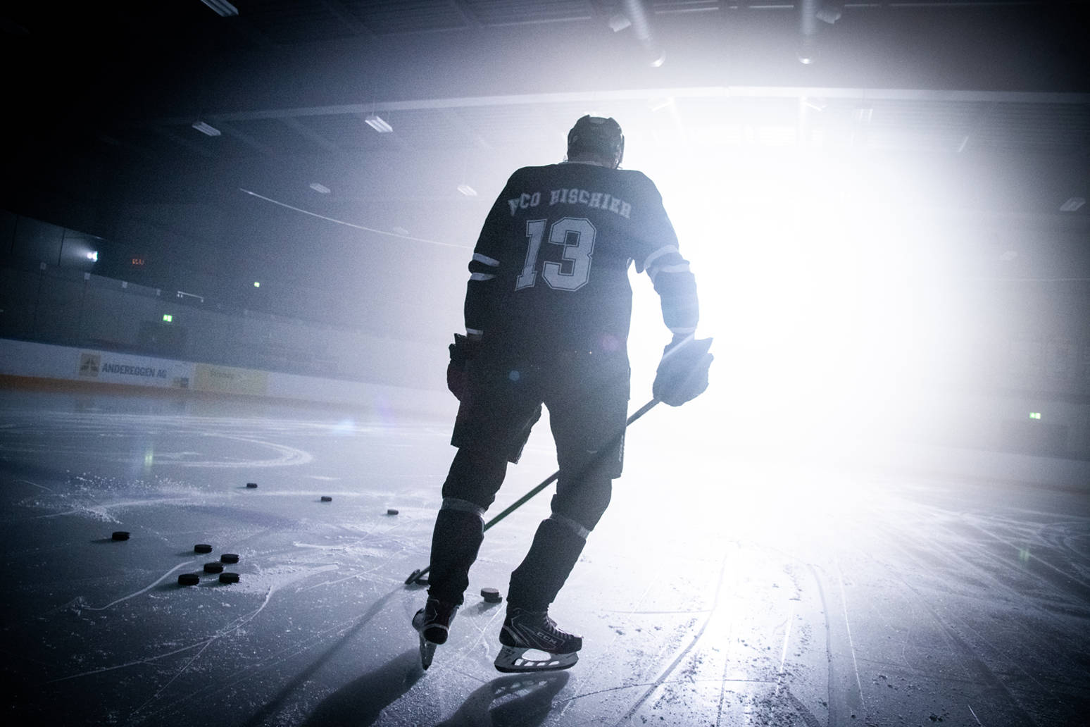 hockey 桌布 壁紙 — 一名身穿13號球衣的冰上曲棍球運動員獨自站在冰場上，被明亮的聚光燈從背後照亮，投下長長的影子。
