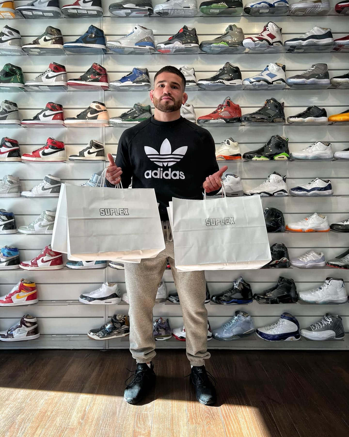 鞋子 桌布 壁紙 — 一名身穿adidas連帽衫的男子，手持兩個Suplex包包，站在一面掛滿色彩繽紛運動鞋的牆前。
