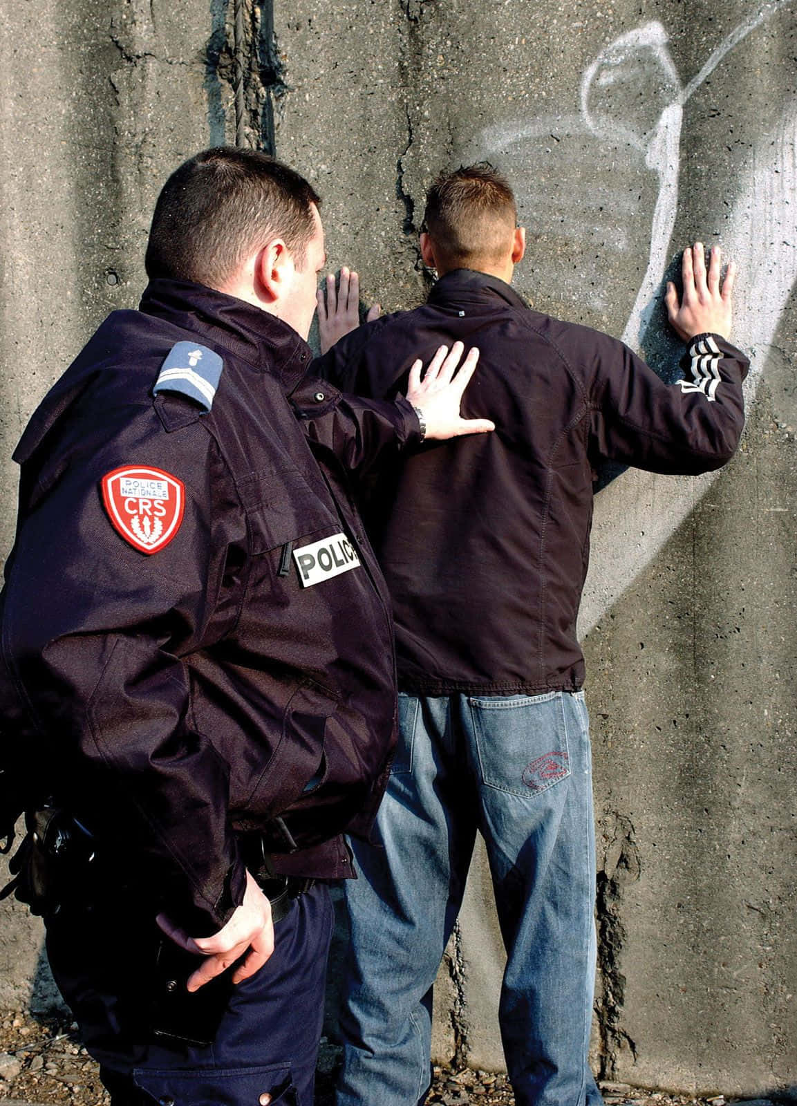 警察 桌布 壁紙 — 一名身穿CRS制服的警察將一名男子按在裂開的混凝土牆上，男子雙手壓在塗鴉覆蓋的表面。